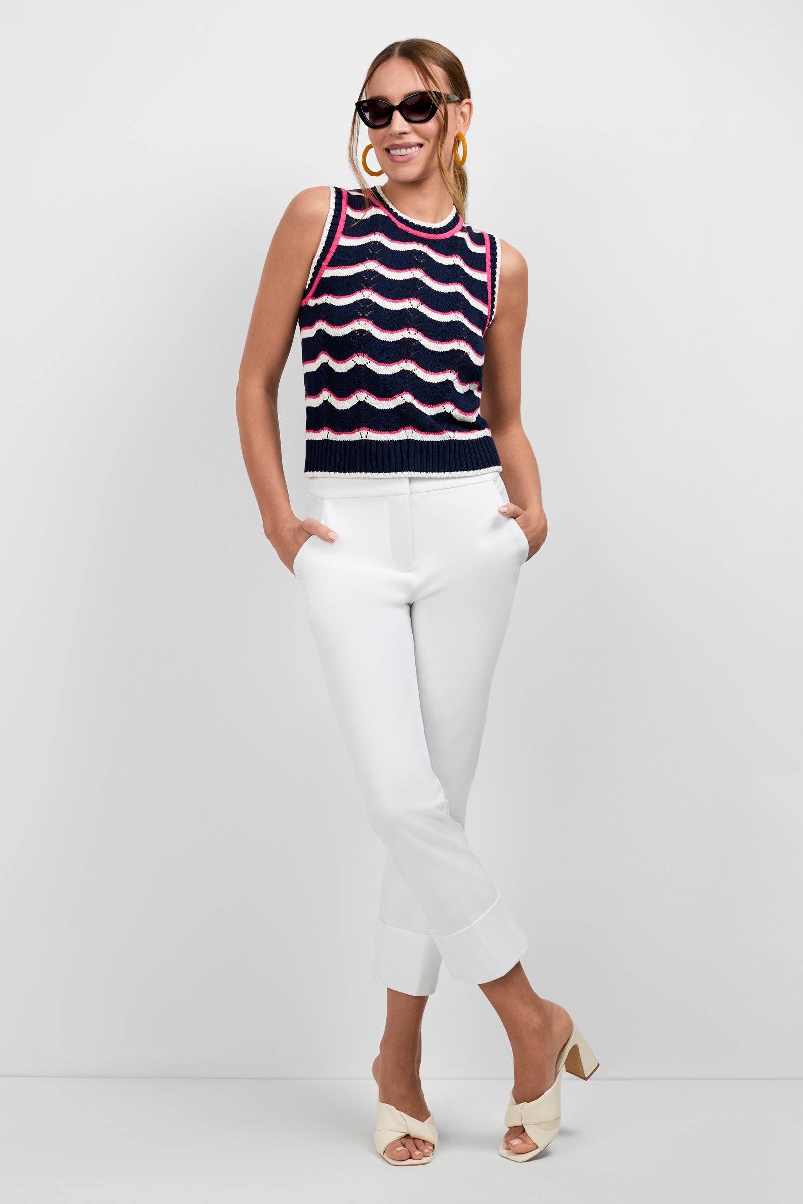A smiling woman stands against a white background with her hands in her pockets, wearing sunglasses, hoop earrings, white cropped pants, beige heeled sandals, and the Cawley Knit Tank featuring navy blue, white, and pink wavy patterns.