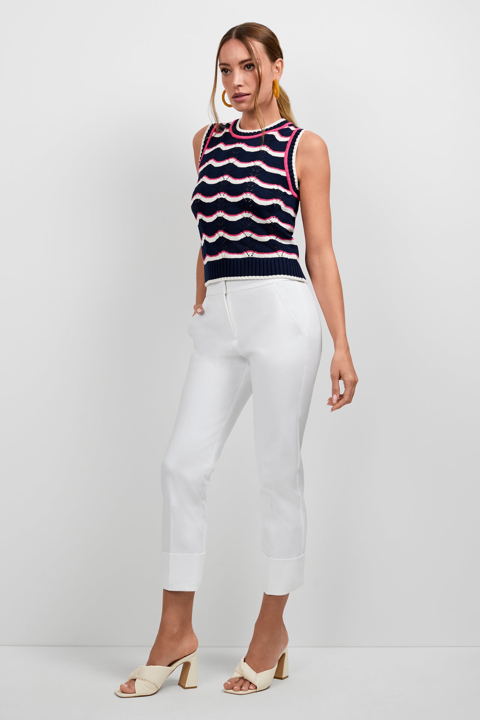 A woman with her hair in a low ponytail stands against a white backdrop wearing the Cawley Knit Tank—a sleeveless, navy, red, and white wavy-striped top—paired with white cropped trousers and cream heeled sandals.