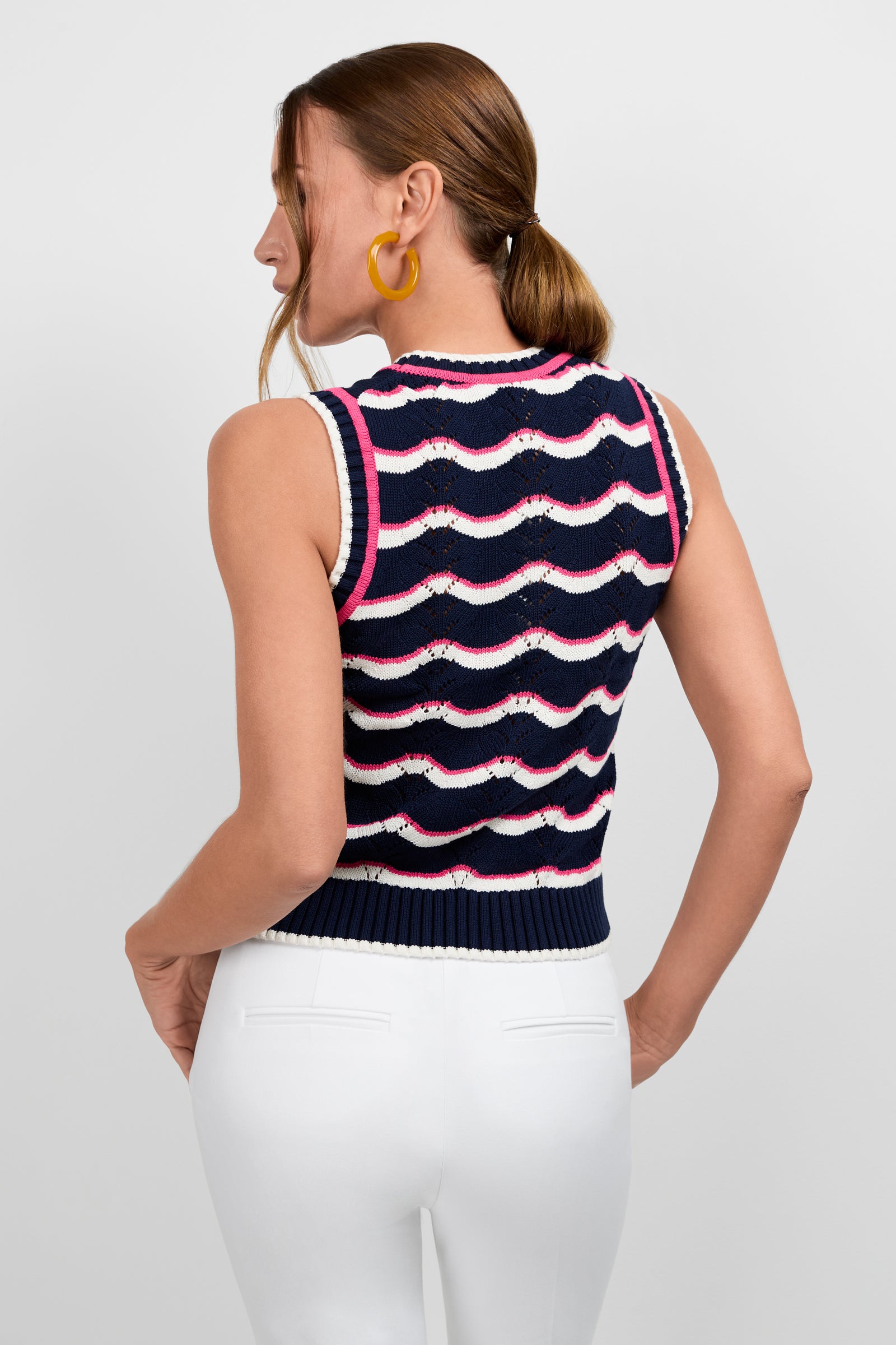 Woman with brown hair in a low ponytail wears the Cawley Knit Tank, featuring navy blue, white, and pink stripes, paired with white pants. She stands with her back turned and slightly to the side, accessorized with large yellow hoop earrings.
