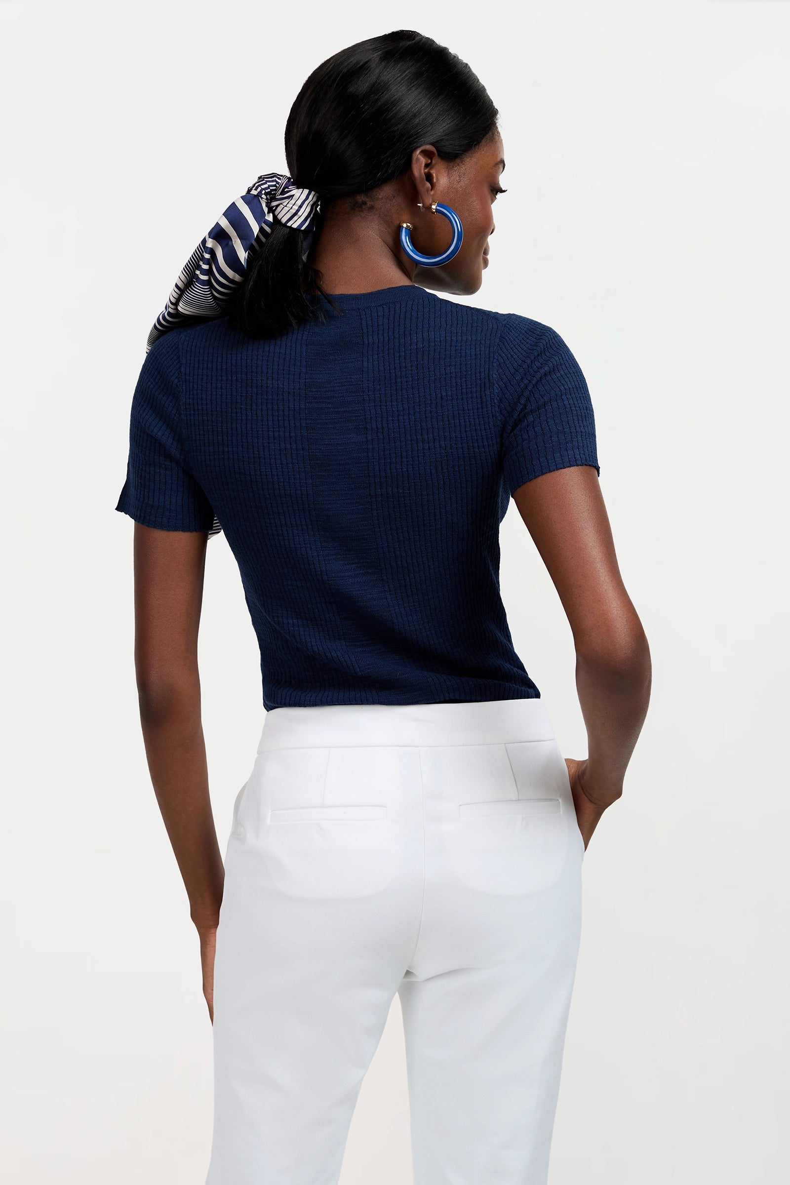 A woman faces away, wearing the Mina Lace-Up Top in navy, white pants, blue hoop earrings, and a striped scarf in her dark hair. She stands against a plain white background.