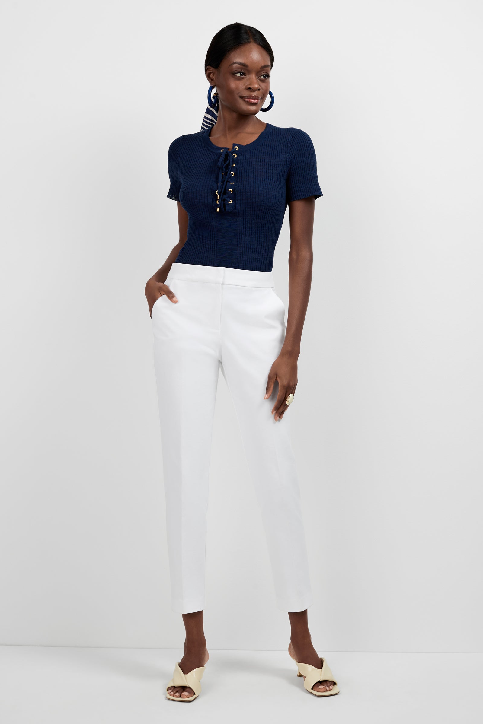 A woman poses against a plain background wearing the Mina Lace-Up Top in navy, paired with white high-waisted pants, beige heeled sandals, and large blue circular earrings. She stands with her hands in her pockets.