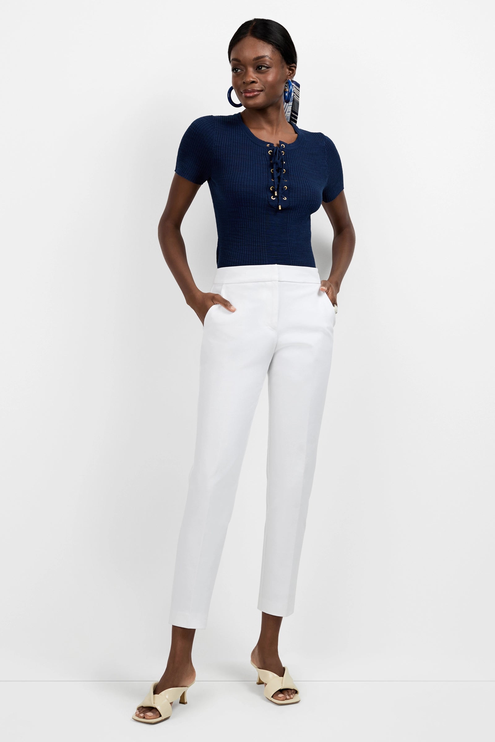 A woman stands against a white background, wearing a navy short-sleeve knit top with front lace-up detail, Aubree 2 Cigarette Pants in white, cream open-toe heeled sandals, and large statement earrings.