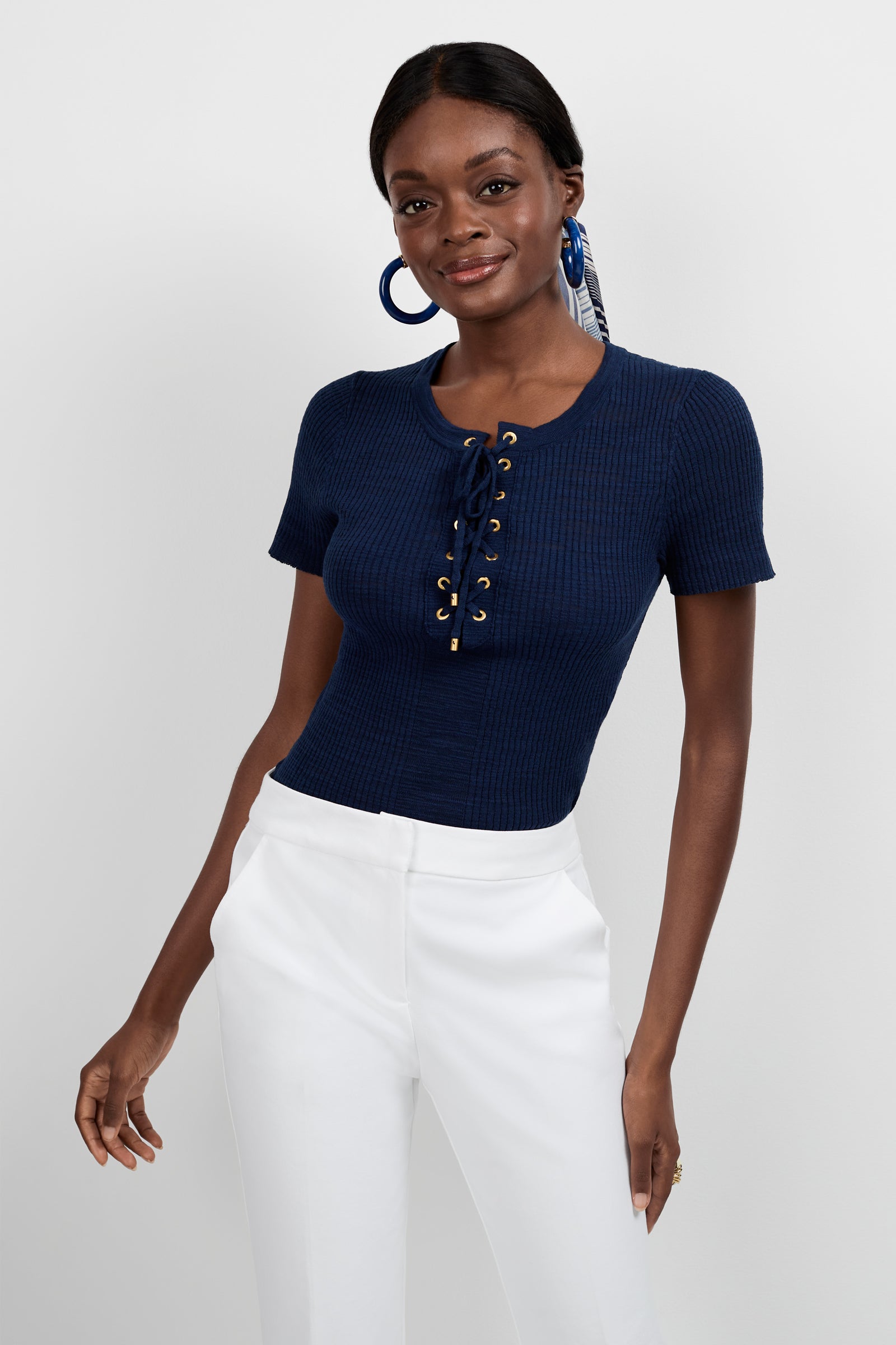 A woman wears the Mina Lace-Up Top, a fitted short-sleeve navy blue top with gold eyelets, paired with high-waisted white pants. She accessorizes with hoop earrings and a patterned hair scarf, smiling against a plain light background.