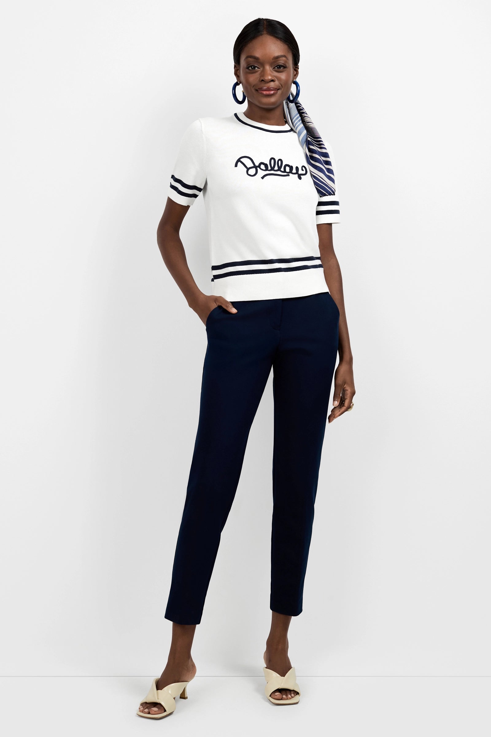 A woman wearing a white short-sleeve sweater with Bally written on it, Aubree 2 Cigarette Pants in navy blue, cream sandals, blue hoop earrings, and a striped scarf tied in her hair stands against a plain white background.