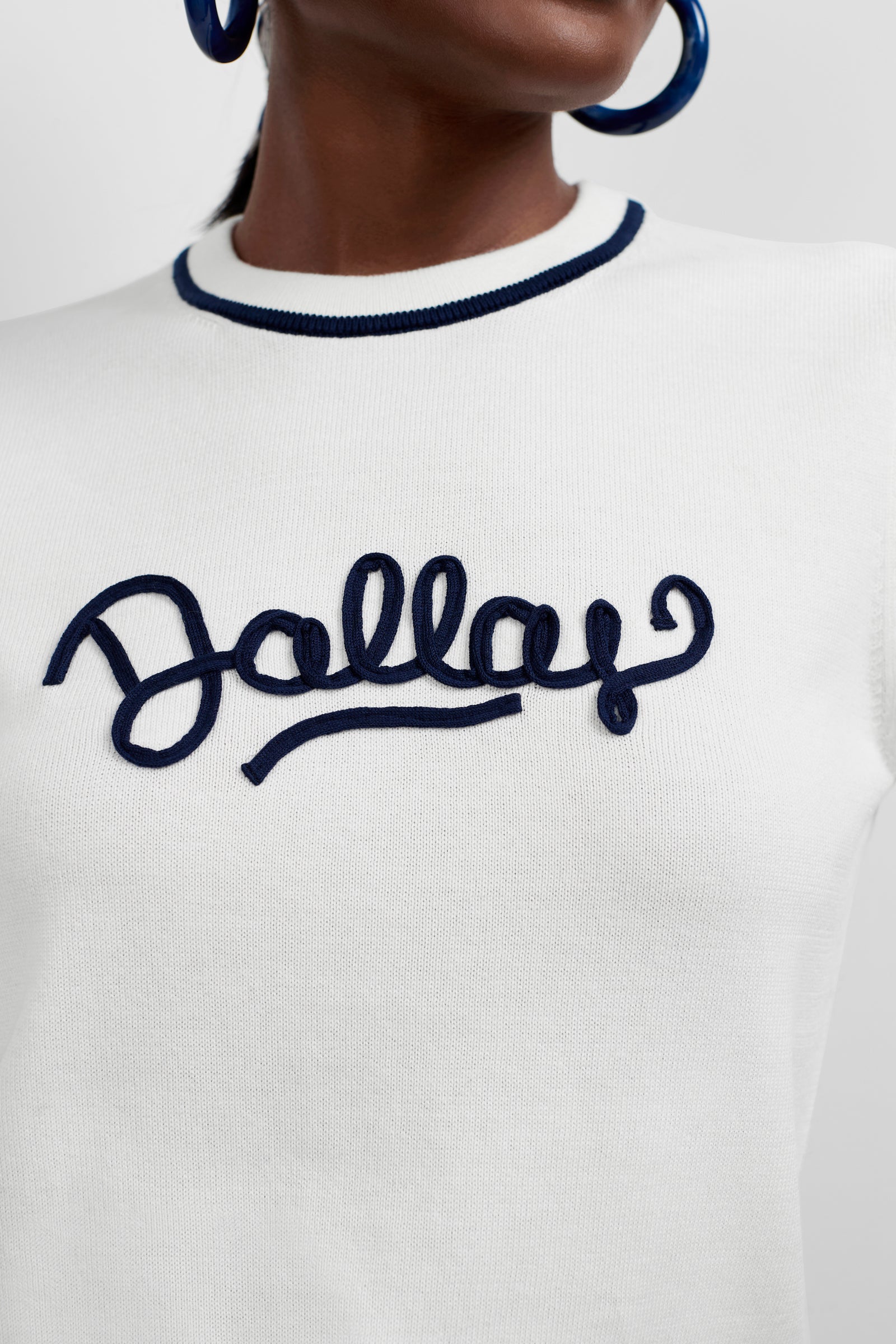 Close-up of a person wearing the Old Fashioned Sweater - Dallas, featuring a white base, dark blue collar trim, and bold blue script lettering spelling Dallas across the chest. The person also sports large blue hoop earrings.