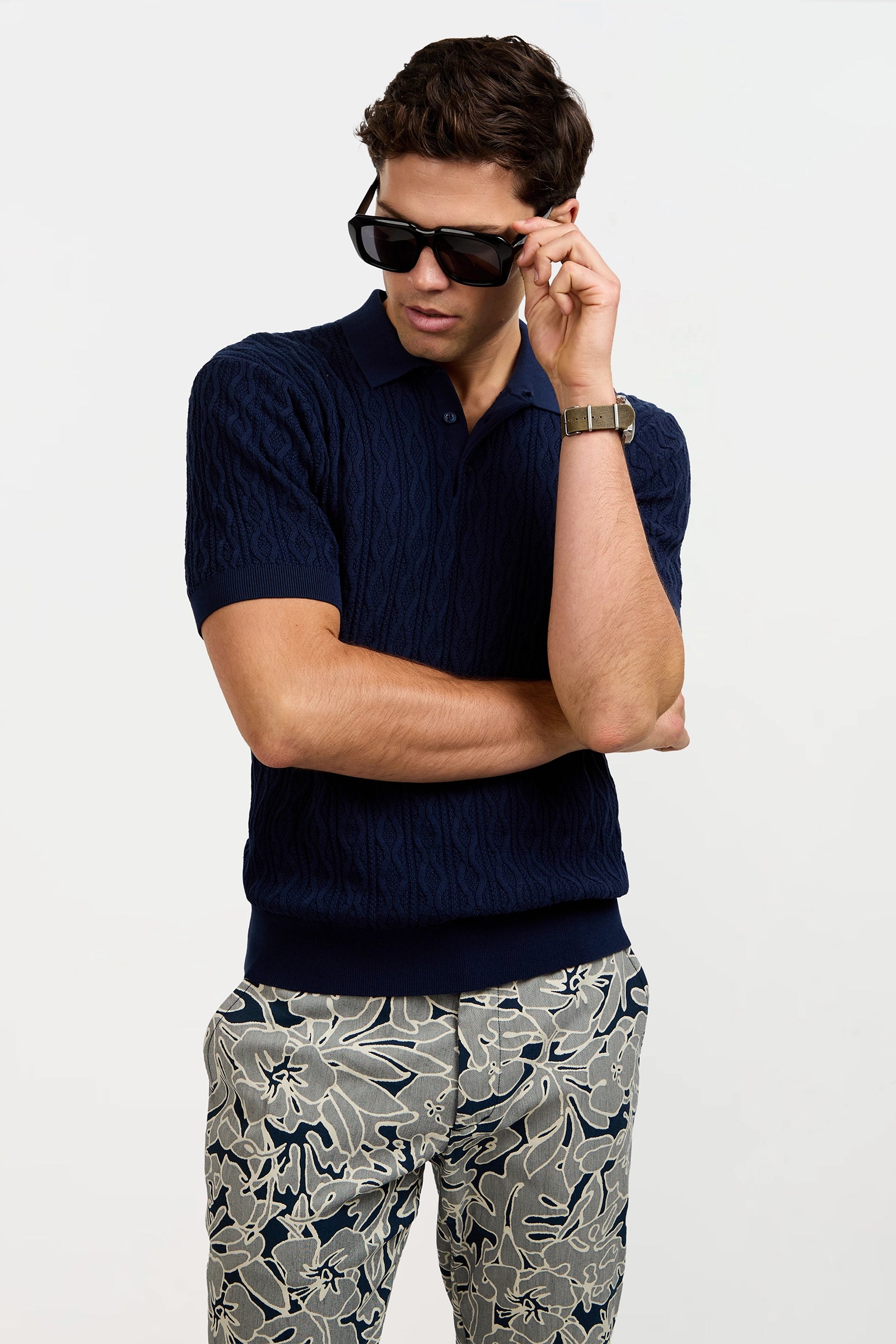 A man wearing sunglasses, gray floral-patterned pants, and the Elio Short-Sleeve Polo in navy stands with one arm crossed and the other adjusting his sunglasses, looking down and to the side.