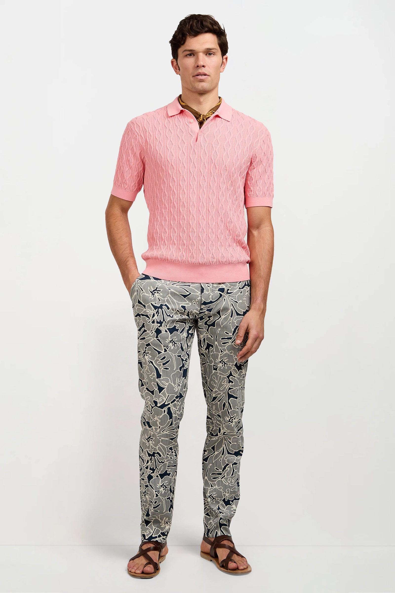A man stands against a white background wearing the Elio Short-Sleeve Polo in light pink, floral slim-fit pants, brown sandals, and a necklace.