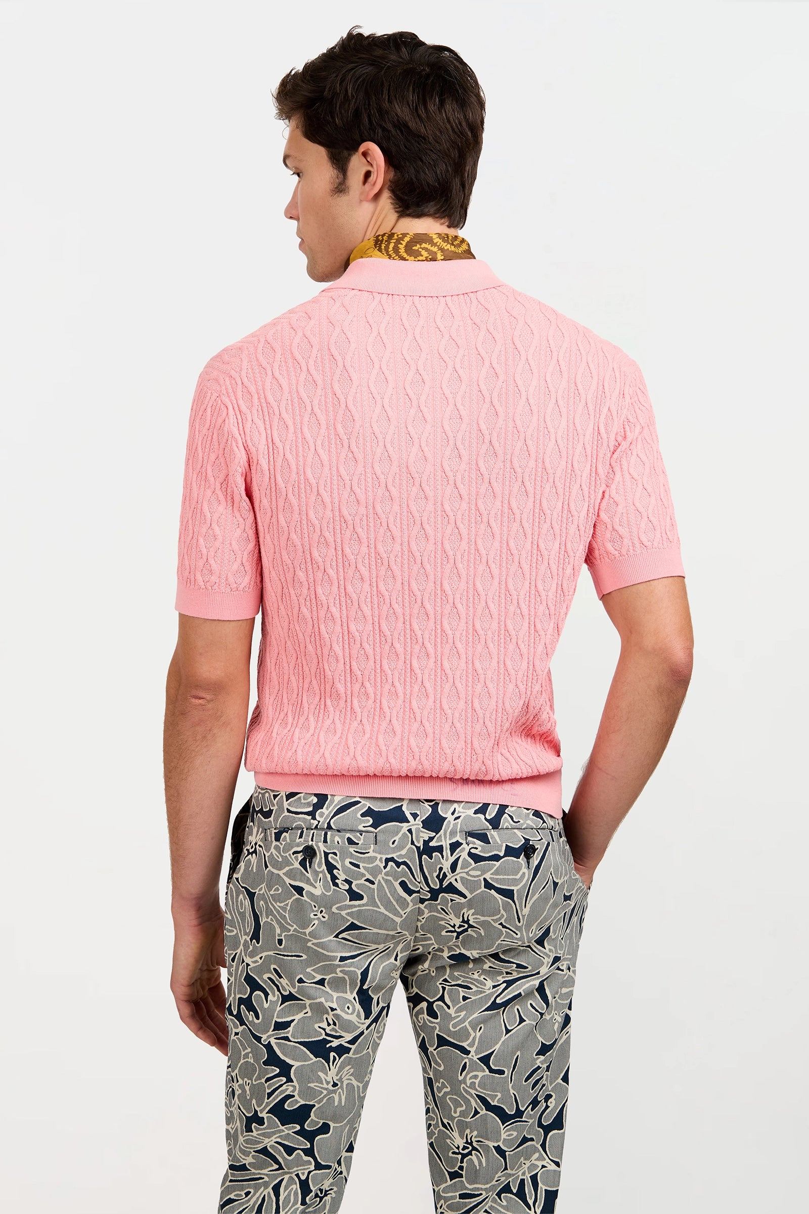 A man stands with his back turned, wearing the Elio Short-Sleeve Polo in light pink, paired with a decorative scarf and blue pants featuring a white floral pattern against a plain, light background.