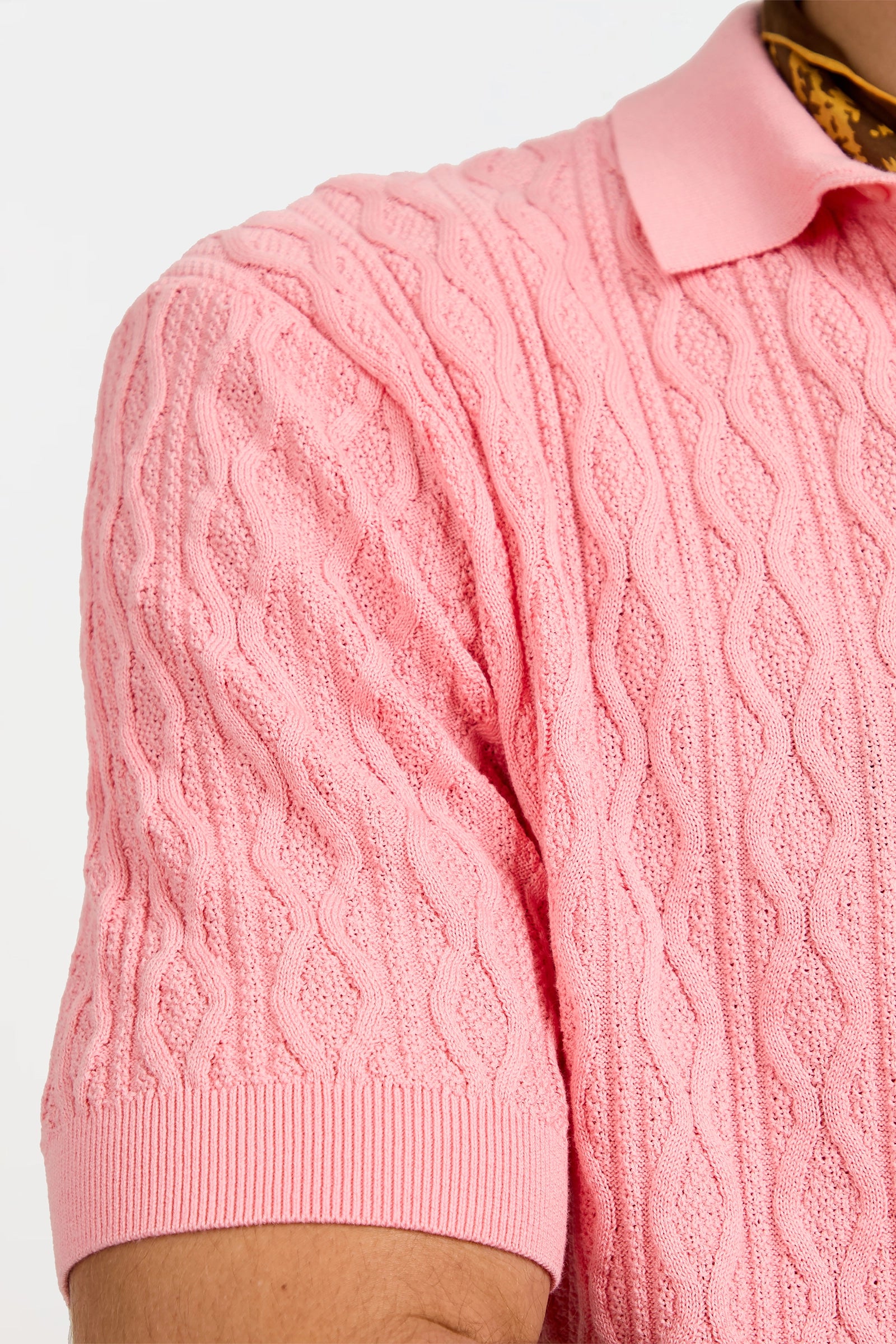Close-up of a person wearing the Elio Short-Sleeve Polo in light pink, featuring cable-knit texture and ribbed cuff and collar. Only the upper arm, chest, and collar area are visible.