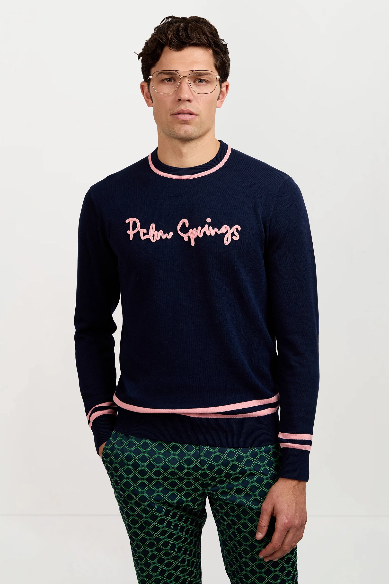 A man in clear glasses and green patterned pants wears the Palm Springs Crewneck Sweater, featuring “Palm Springs” in pink on navy, while standing against a plain light background.