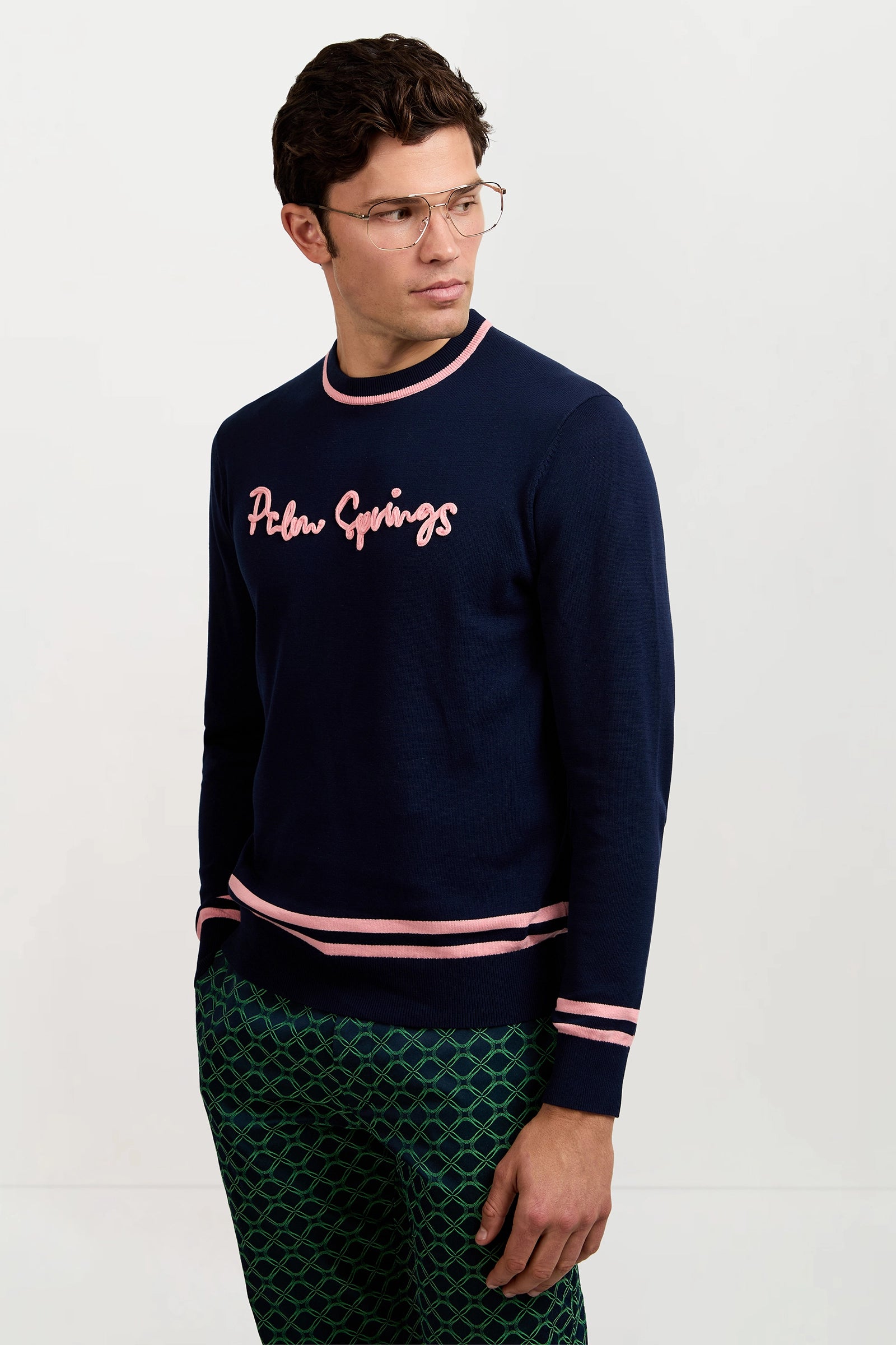 A man wearing glasses and the Palm Springs Crewneck Sweater in navy blue with pink script stands against a plain white background, looking slightly to his left.