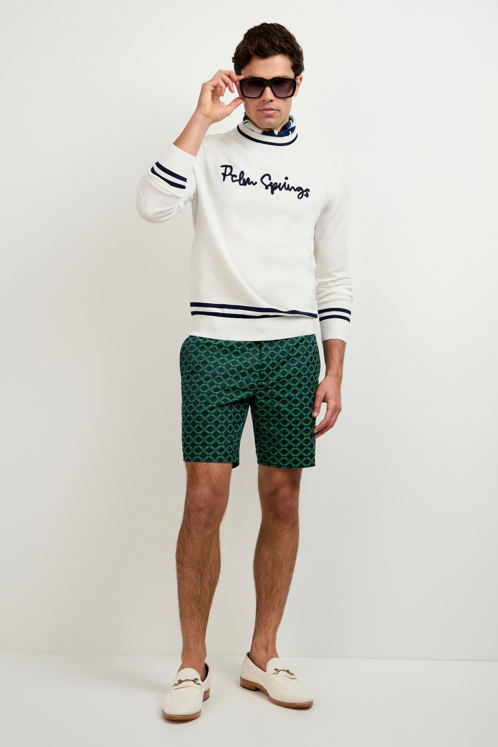 A man poses against a white background wearing dark sunglasses, a striped white Palm Springs sweater, a patterned blue neck scarf, Lawrence Short - Clover Geo Jacquard, and white loafers.