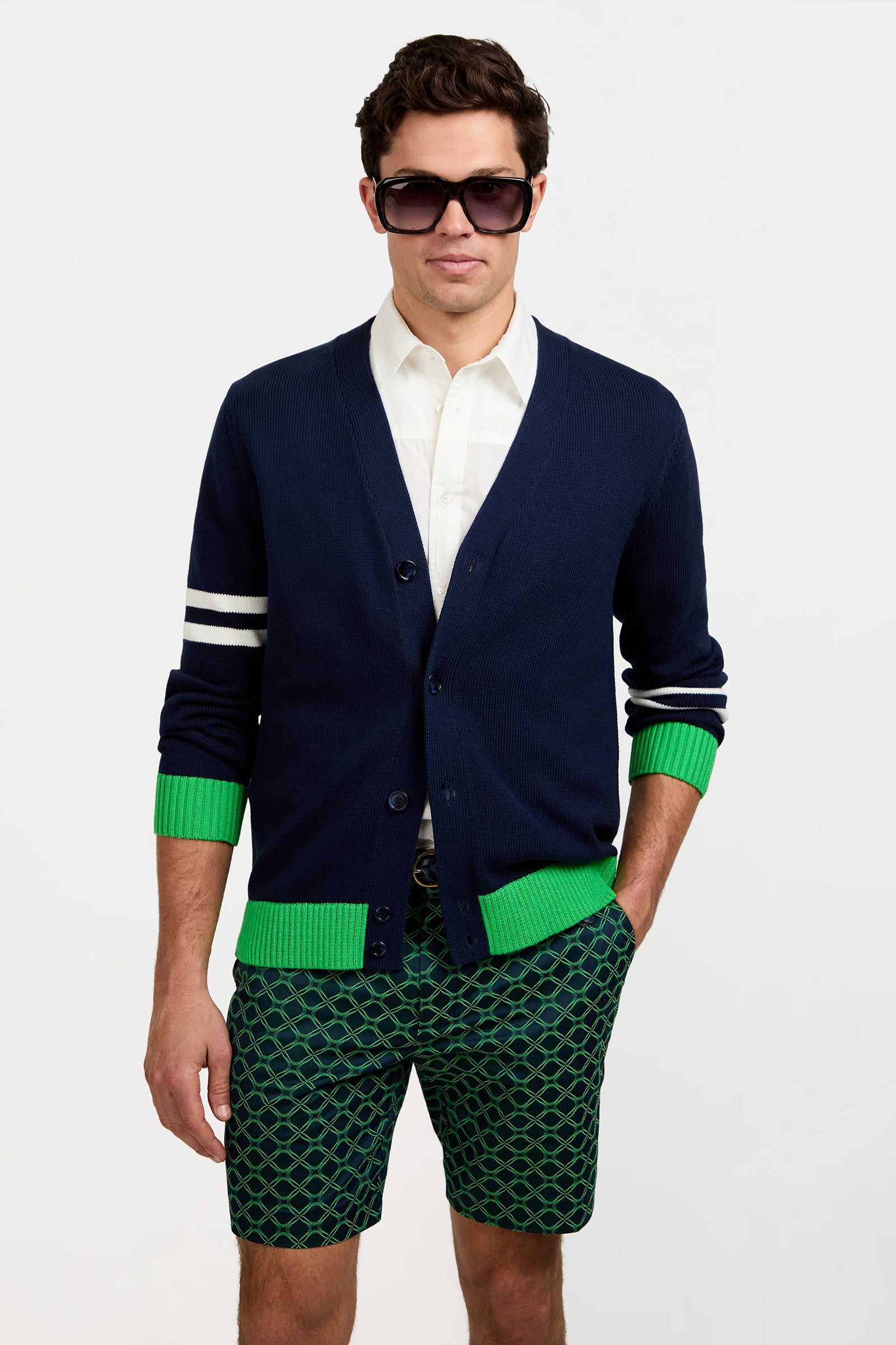 A man wearing dark sunglasses, the Amadeo Cardigan with white stripes and green cuffs, a white shirt, and green patterned shorts stands against a plain white background.
