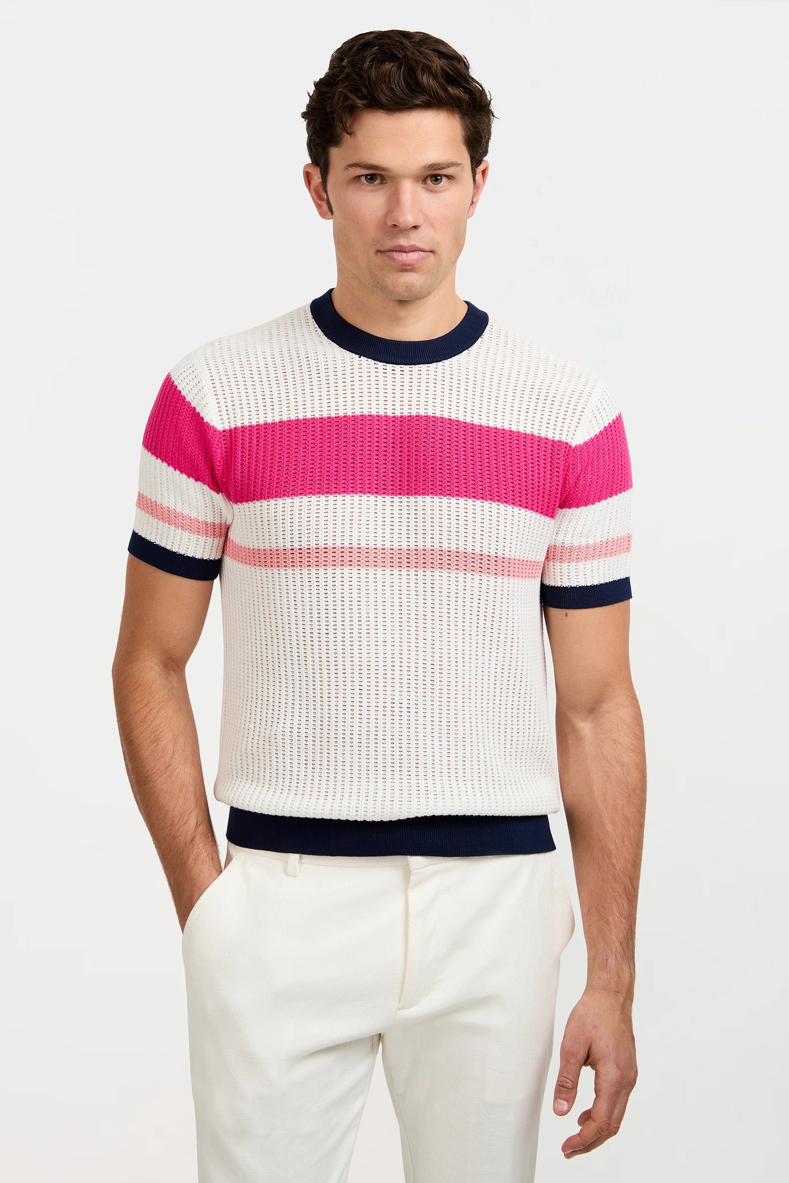 A man wearing the Aldo Short-Sleeve Crewneck with navy, pink, and cream stripes and white trousers stands against a white background, his right hand in his pocket while he looks at the camera.