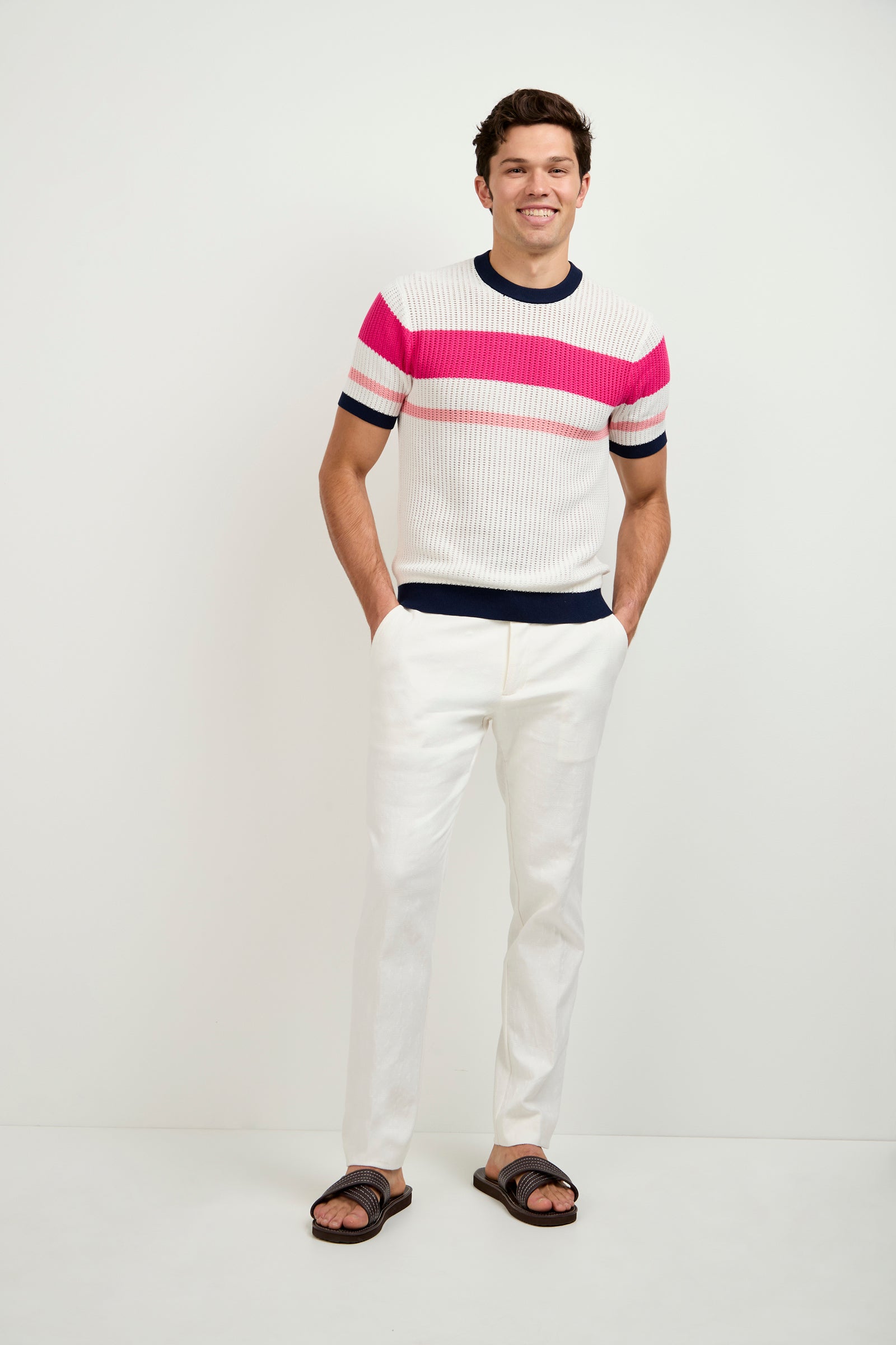 A man stands against a plain white background, smiling with his hands in his pockets, wearing the Aldo Short-Sleeve Henley featuring pink and red horizontal stripes, paired with white pants and brown sandals.