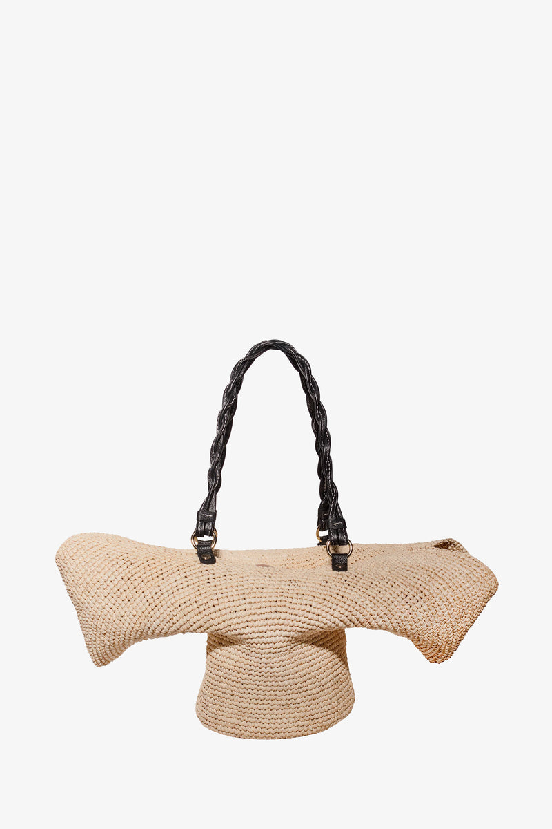 The Hat Tote is a natural-colored woven straw handbag featuring an abstract, sculptural shape and two twisted black handles, shown against a plain white background.