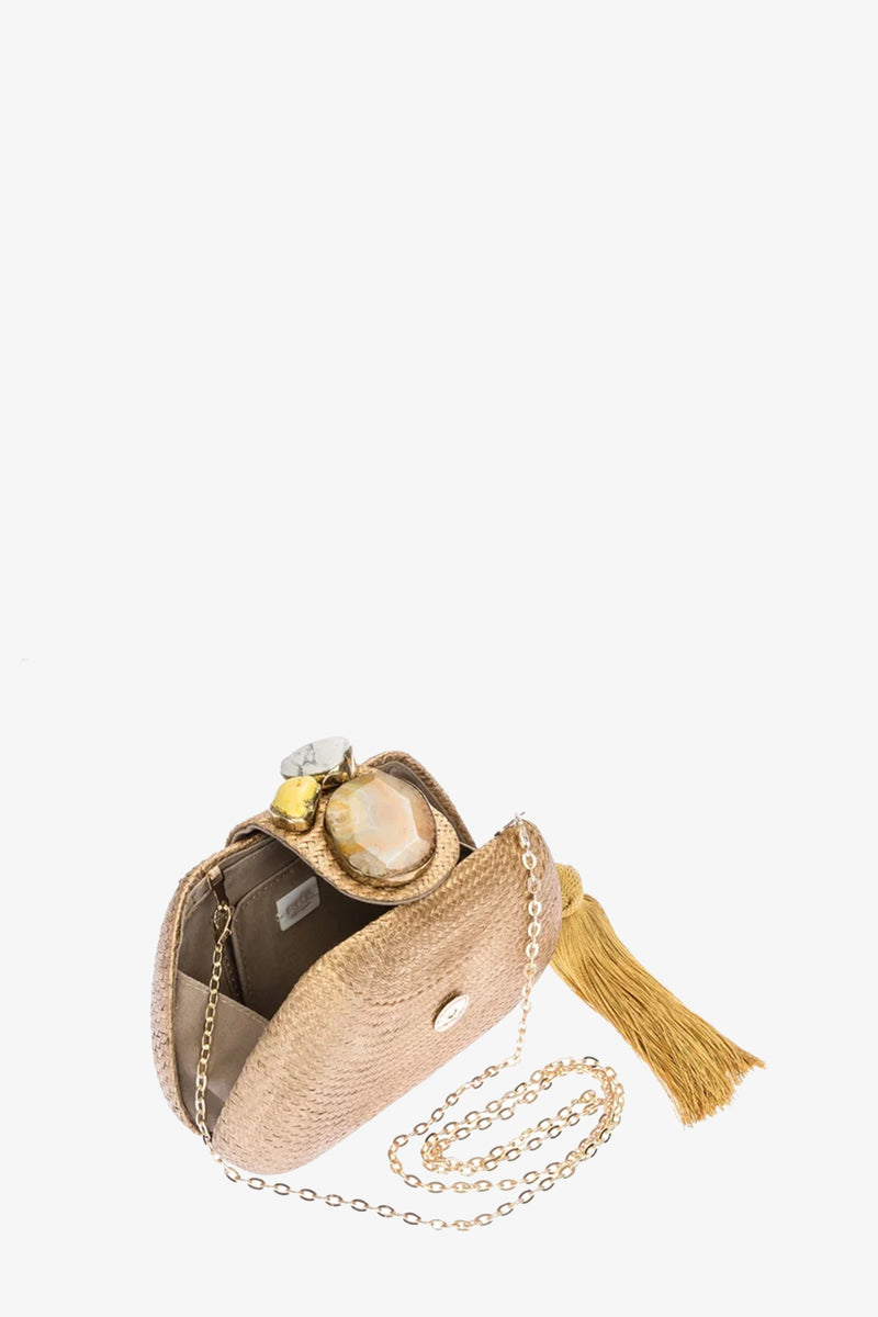 The Tamu Bean Clutch is a small beige woven purse with a gold chain strap, a large yellow tassel on the side, and an open top displaying a perfume bottle inside.