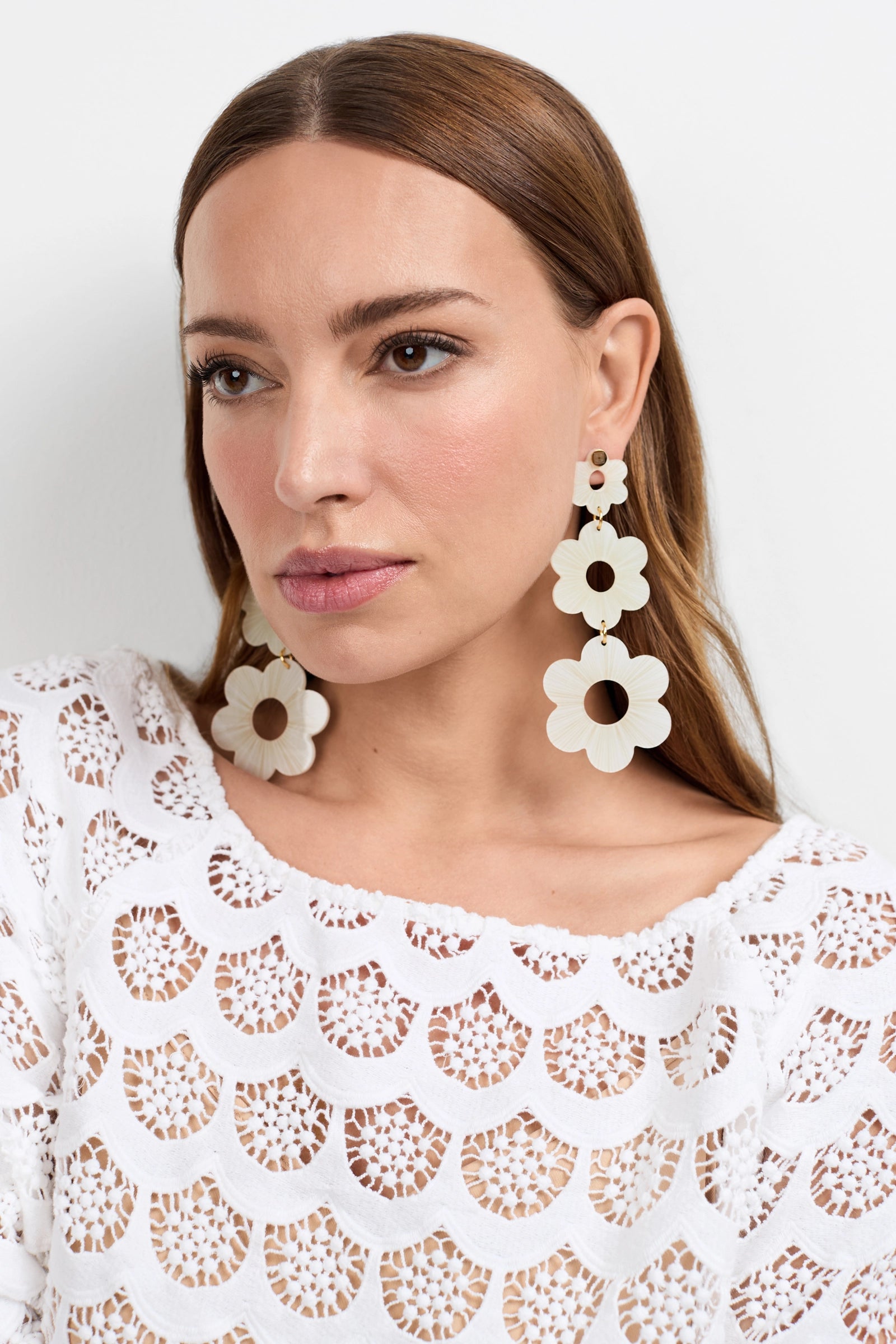 A woman with straight brown hair wears the Sunny Daisy Earrings - Large in white and a white crochet top. She has a neutral expression and stands against a plain, light background.