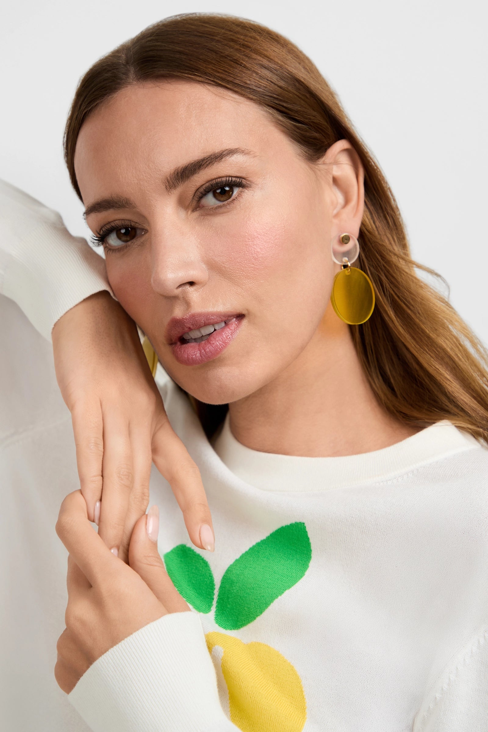 A woman with light brown hair poses in a white sweater with a lemon design, wearing Citron Round Drop Earrings in citron. Her hand is raised near her face, and she gazes just past the camera with a neutral expression.