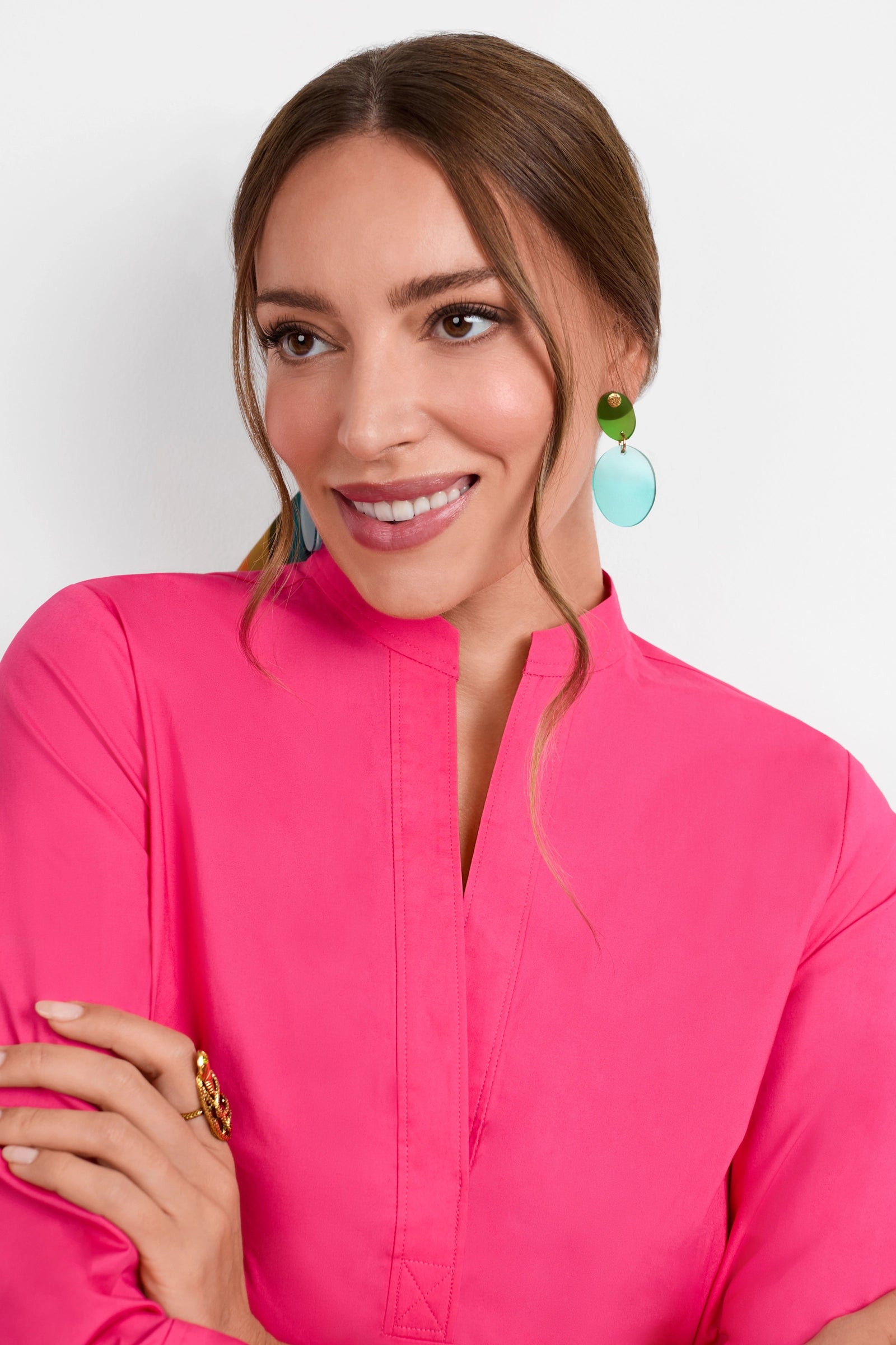 A woman with long brown hair tied back, wearing a bright pink top and Ocean Droplet Earrings, smiles while looking to the side against a plain white background.