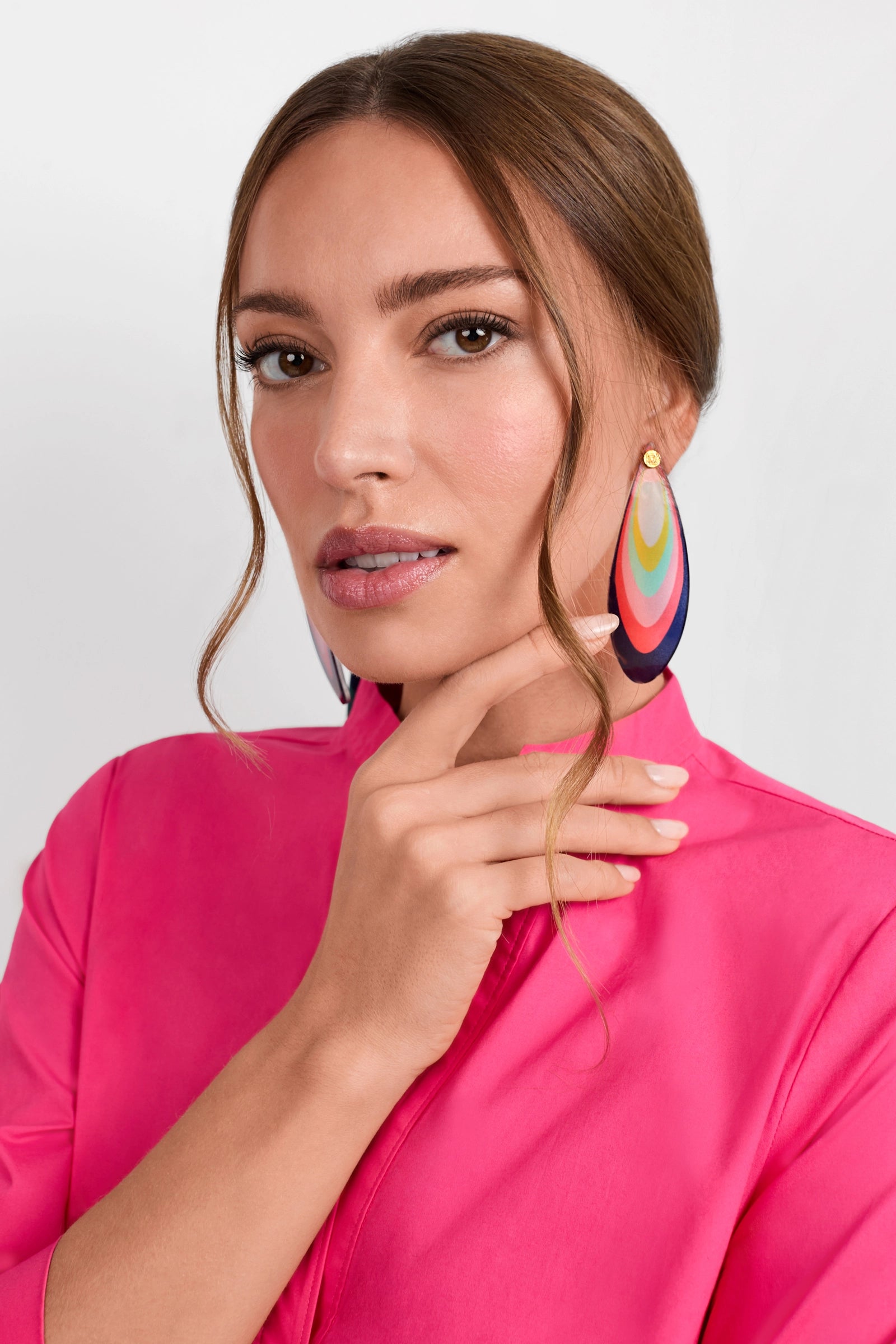 A woman in a bright pink top poses with her hand near her chin, wearing the rainbow Aster Earrings—large and teardrop-shaped. She has light makeup, and her brown hair is styled neatly with a loose strand framing her face.