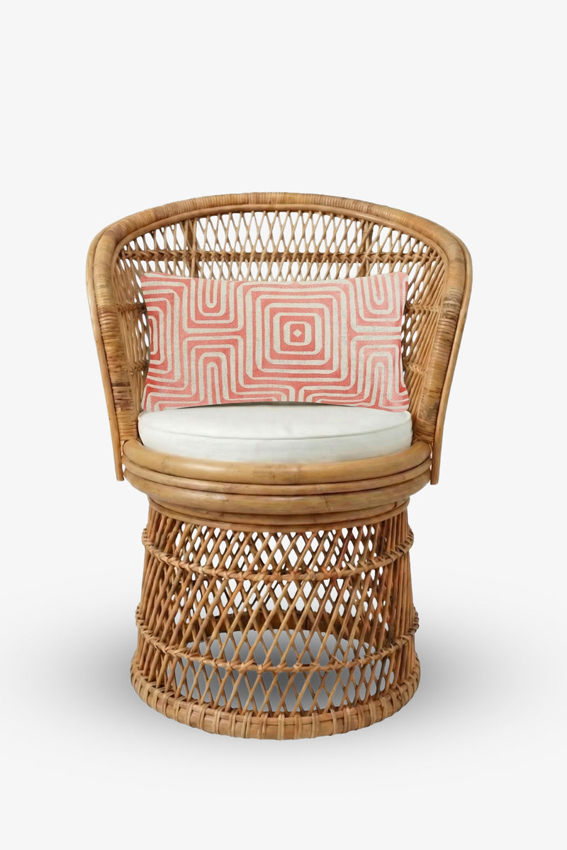 A round wicker chair with a white cushion is styled with the Amazing Maze Embroidered Lumbar Pillow, showcasing its pink and white geometric pattern, all set against a plain white background.