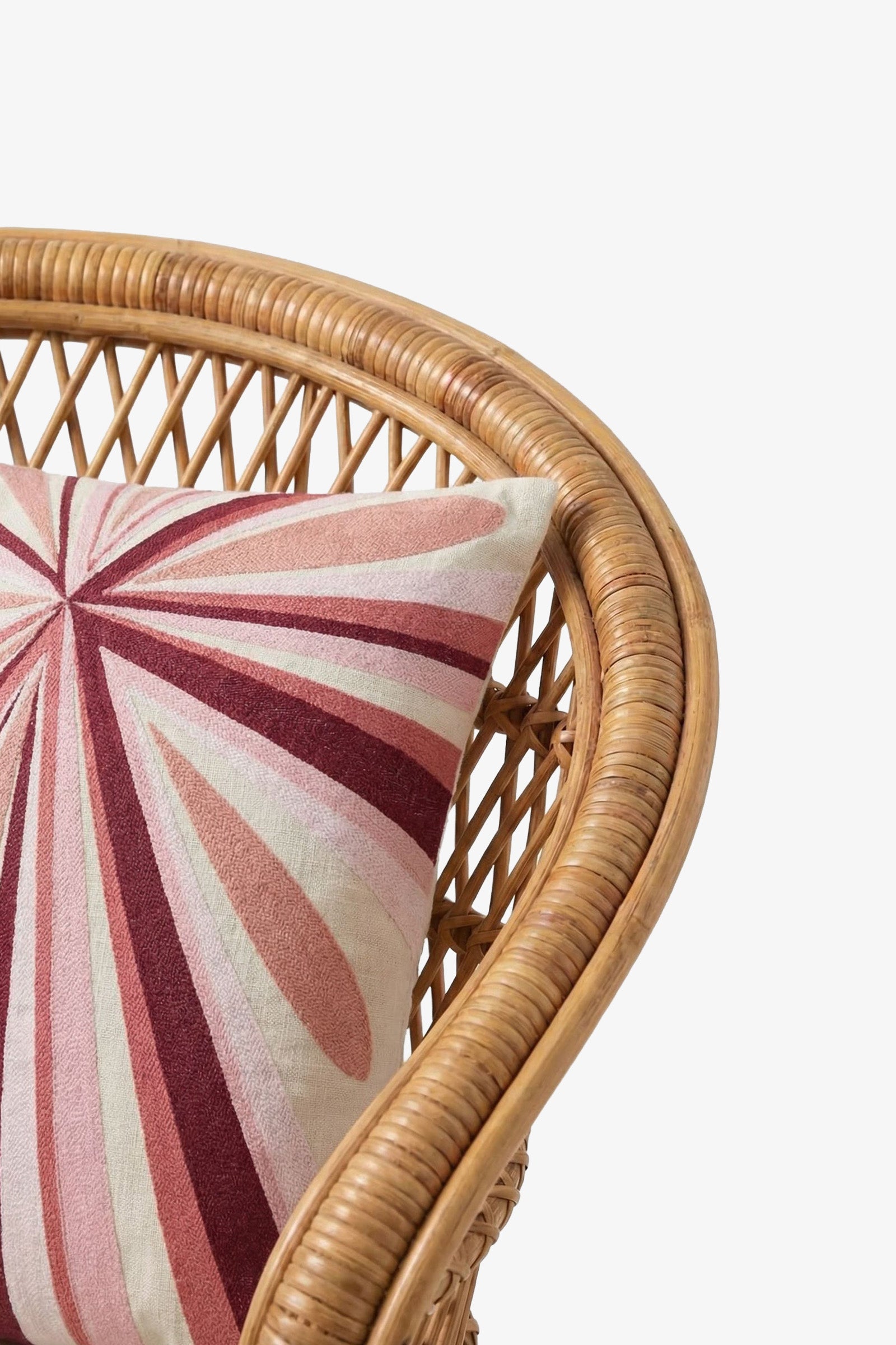 A close-up of a rattan chair featuring the Shante Embroidered Square Pillow, adorned with a pink, red, and cream geometric starburst pattern.