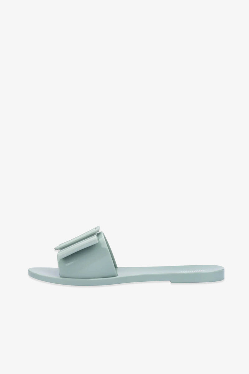 The Babe Sandals are flat slide sandals in light blue, featuring a wide strap adorned with a large decorative bow, displayed against a plain white background.