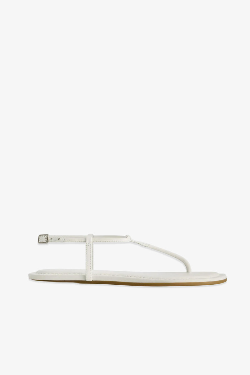 The Hercu Sandal is a minimalist white flat sandal featuring a thin strap across the toes and an ankle strap secured by a small buckle, displayed against a plain white background.