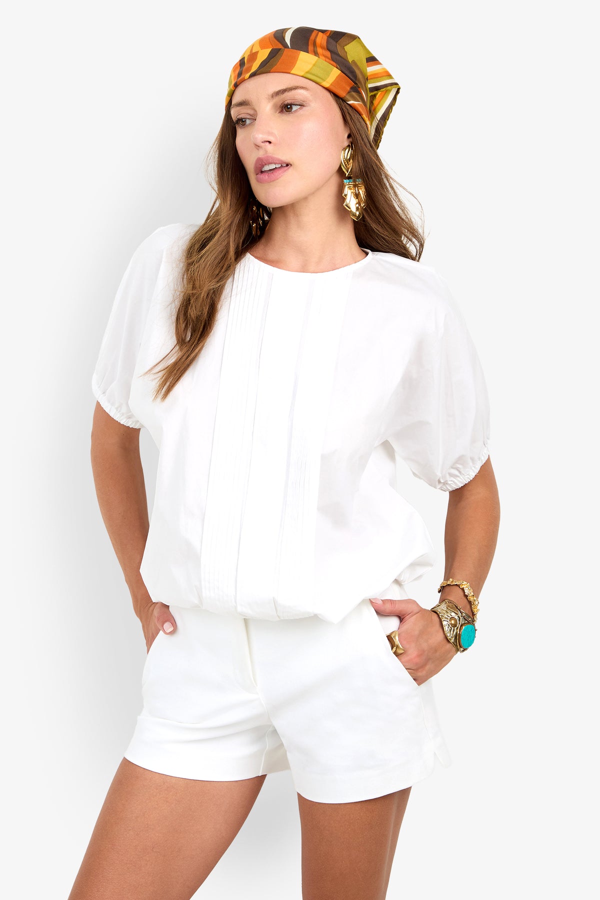 Woman wearing the San Siro Top with matching shorts, colorful patterned scarf, gold earrings, and turquoise jewelry, standing with one hand in her pocket against a white background.