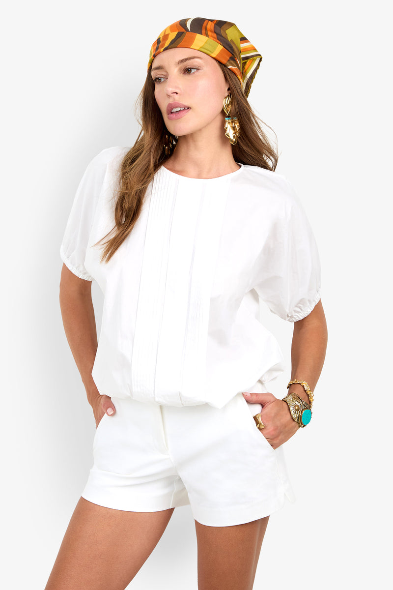 Woman wearing the San Siro Top with matching shorts, colorful patterned scarf, gold earrings, and turquoise jewelry, standing with one hand in her pocket against a white background.
