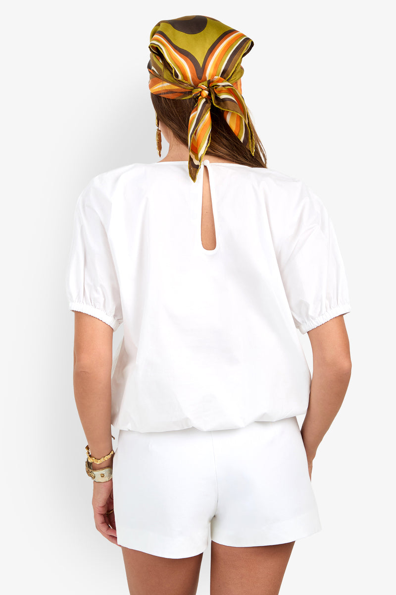 Woman facing away, wearing the San Siro Top in white with matching shorts. She accessorizes with a vibrant patterned scarf and bracelets on her wrist.
