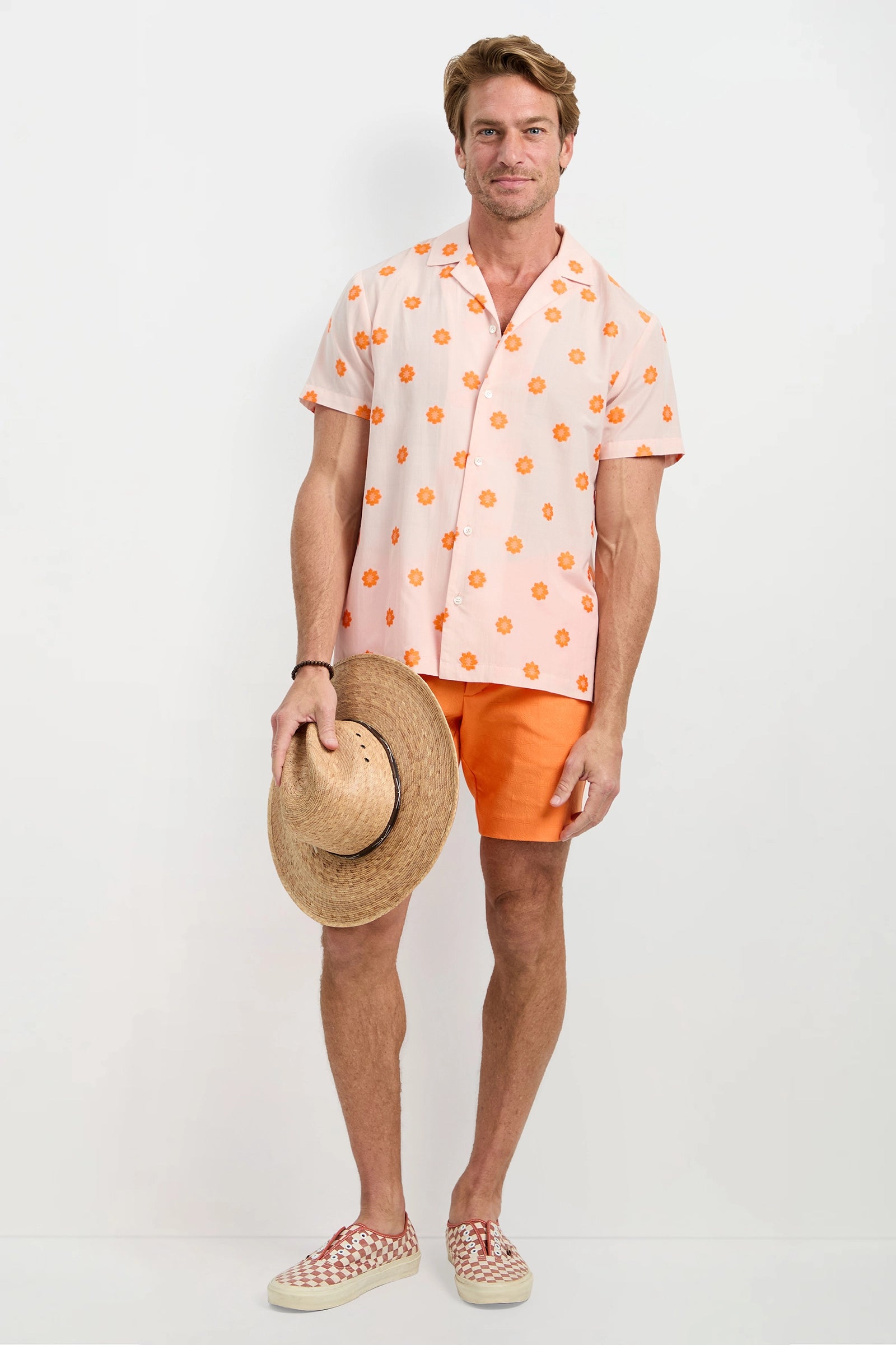A man stands against a white background, wearing the Felix Camp Shirt - Daisy Dots, orange shorts, patterned slip-on shoes, and holding a straw hat with a relaxed expression.