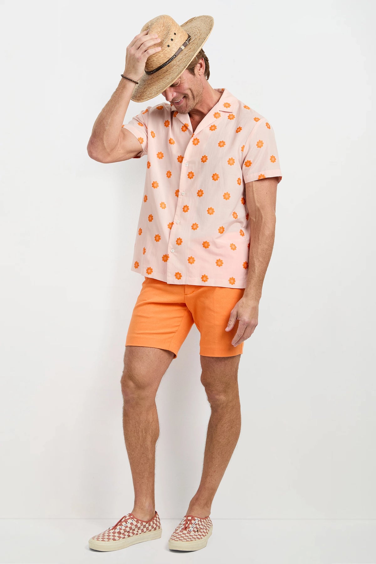 A man wears the Felix Camp Shirt - Daisy Dots, bright orange shorts, checkered slip-on shoes, and a straw hat, smiling and tipping his hat against a plain white background.