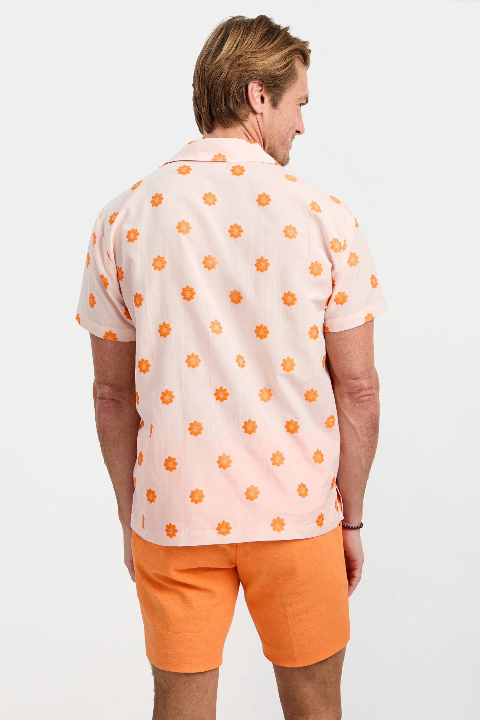 A person with light brown hair faces away, wearing the Felix Camp Shirt - Daisy Dots, an off-white short-sleeve shirt with an orange floral pattern and matching orange shorts against a plain white background.