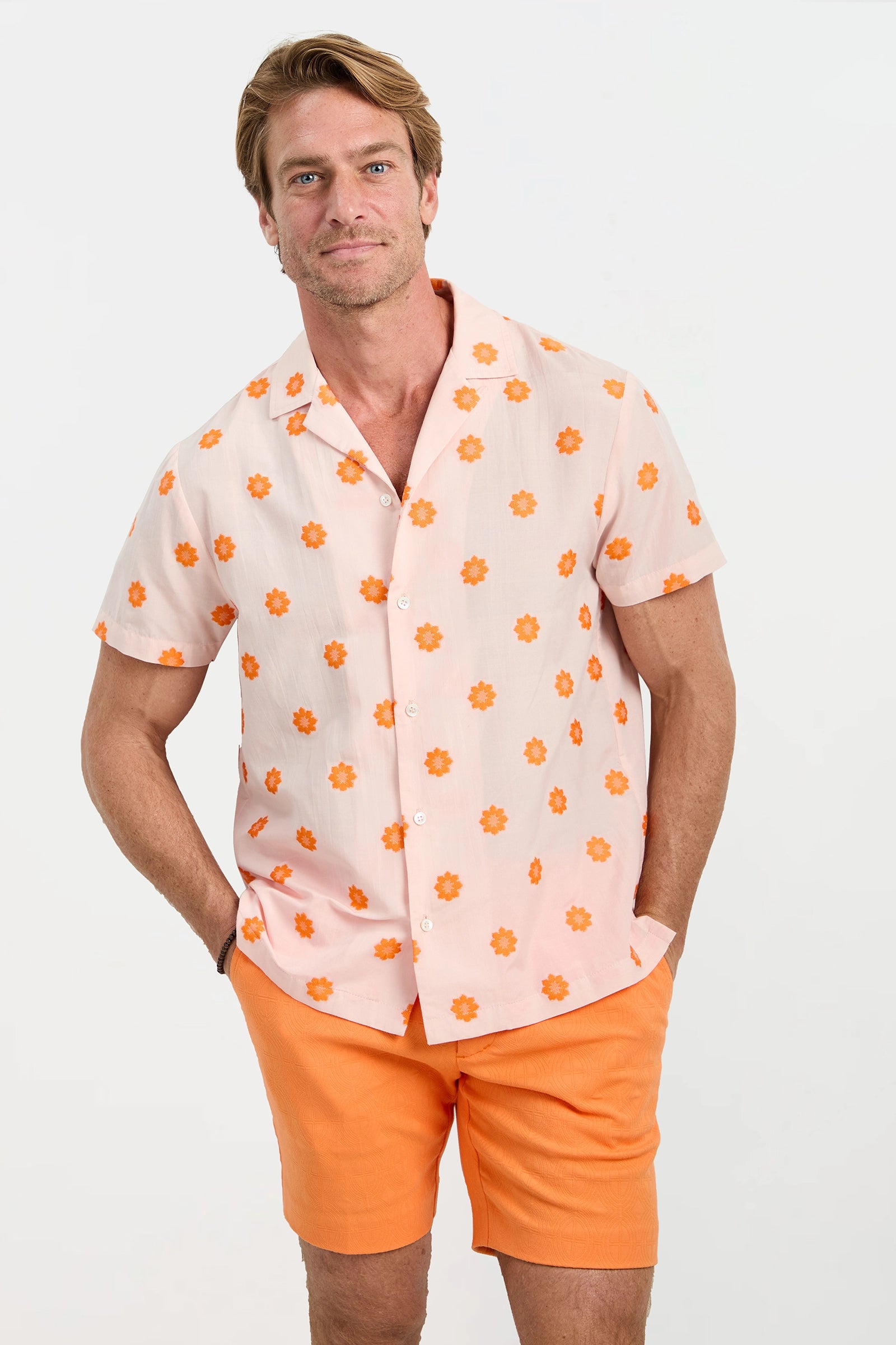 A man stands against a white background wearing the Felix Camp Shirt - Daisy Dots, a light pink short-sleeve shirt with orange floral patterns, paired with orange shorts. He has one hand in his pocket and is smiling.