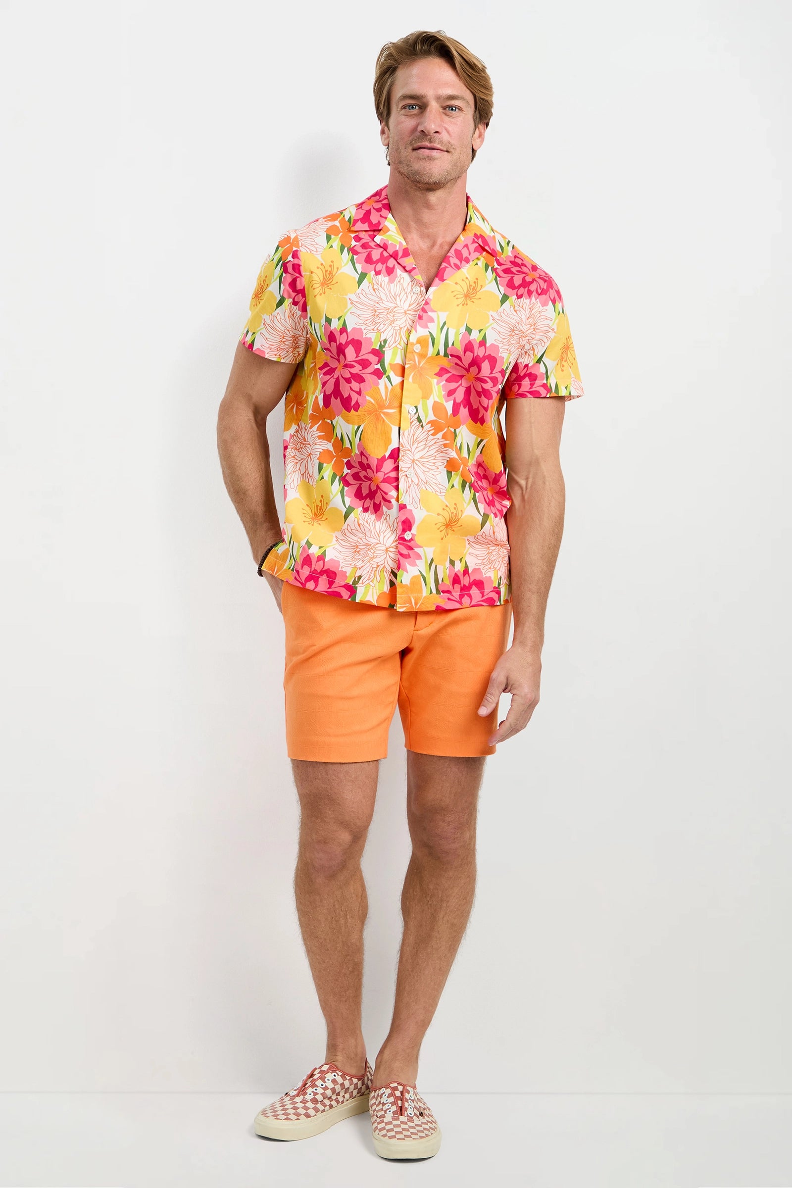 A man stands against a white background, wearing the Felix Camp Shirt - Como, featuring a vibrant pink, yellow, and orange floral pattern. He pairs it with orange shorts and red-and-white checkered slip-on shoes.