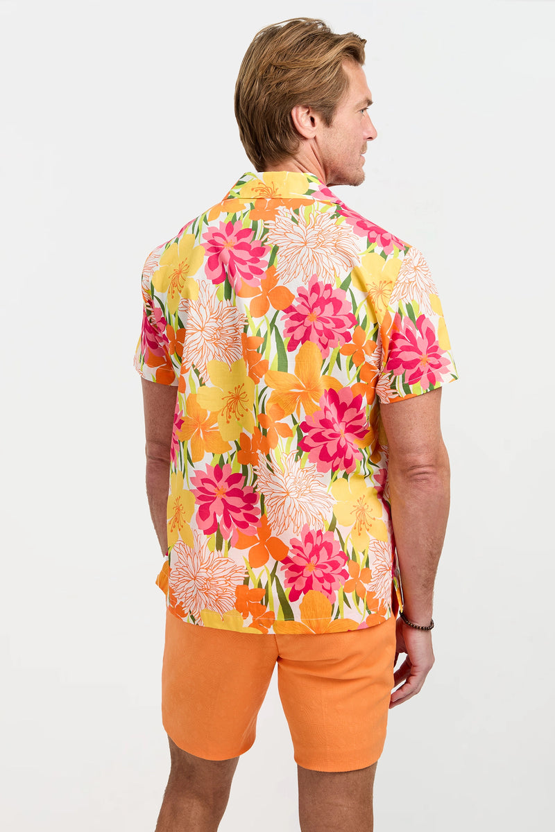 A man with light brown hair wears the Felix Camp Shirt - Como, a bright, short-sleeve button-up with a colorful floral print in pink, orange, and yellow, paired with orange shorts. He stands facing away from the camera.