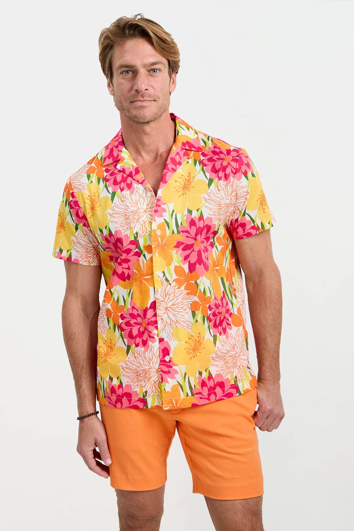 A man stands against a plain background wearing the Felix Camp Shirt - Como, a short-sleeve button-up with a bright floral pattern in yellow, pink, red, and white, paired with solid orange shorts.