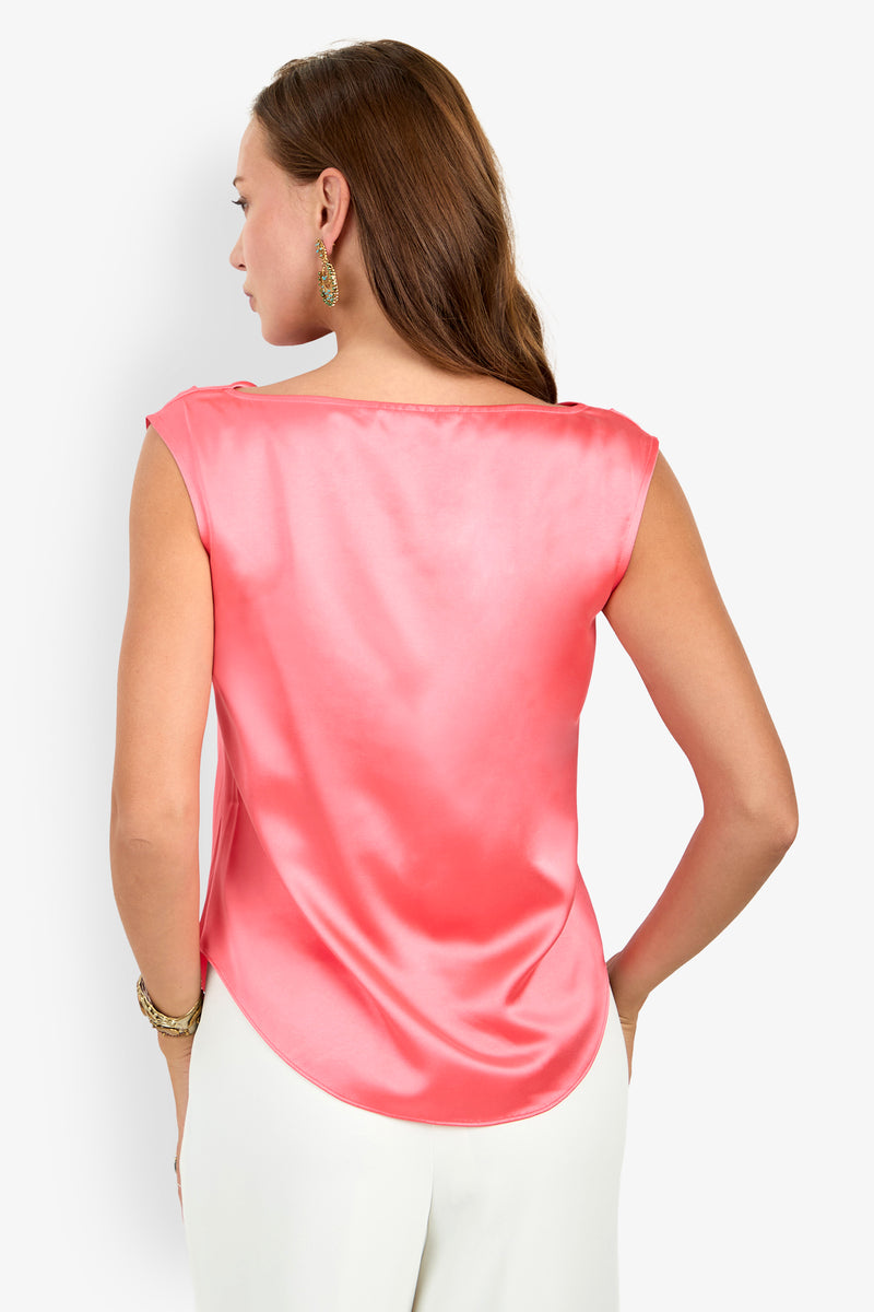 A woman with long brown hair wears gold earrings and the Alessi Cowl Neck Top (Wholesale)—a shiny pink sleeveless blouse—paired with white pants and gold bracelets, standing against a plain white background.