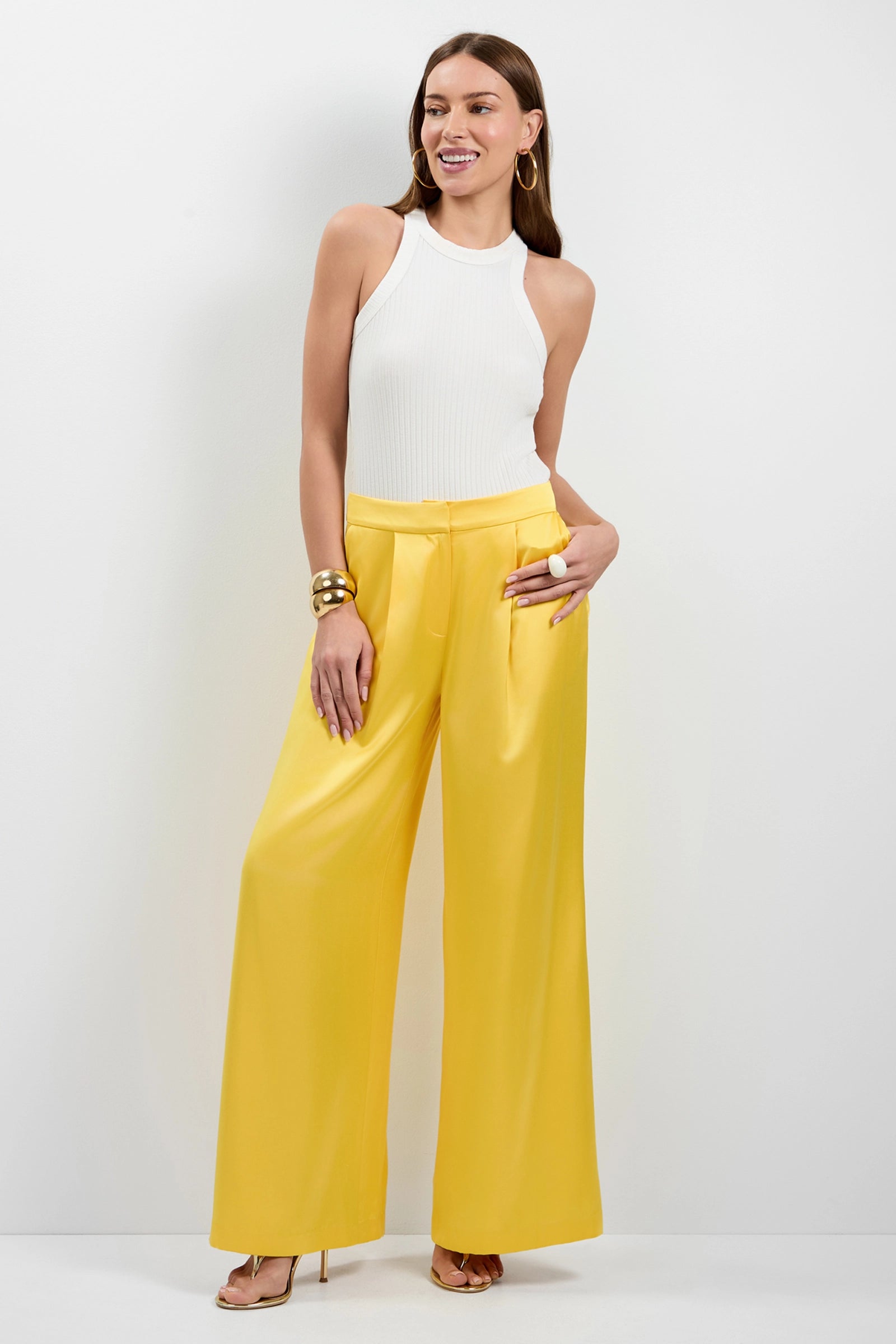 A woman smiles confidently in a sleeveless white ribbed top and Pontaccio Satin Pants in Lambretta Lemon, accessorized with gold hoop earrings, chunky gold bracelets, and gold heels, standing against a white background.