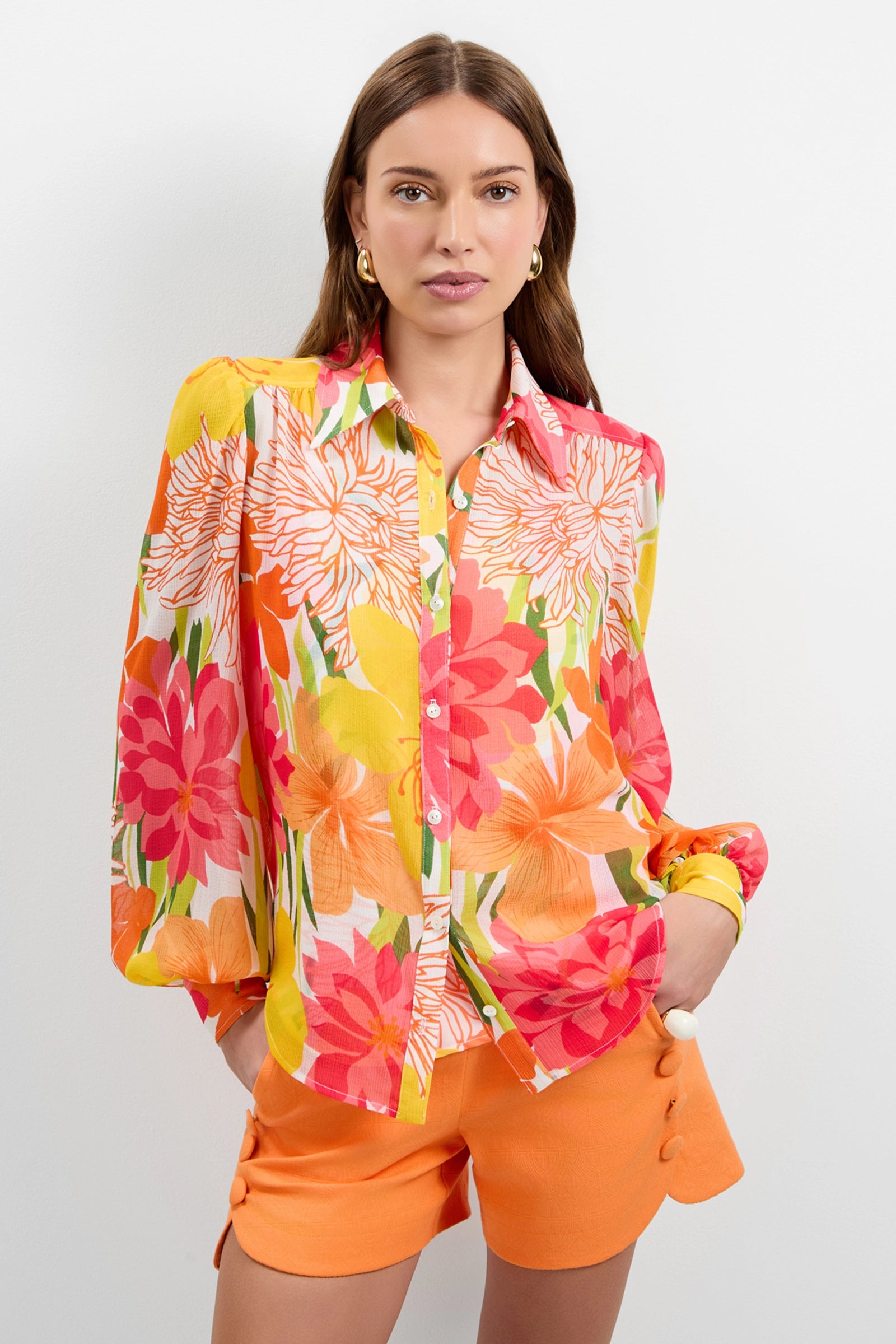 A woman with long brown hair and gold hoop earrings stands against a white background wearing the Monte Balloon Sleeve Top, a colorful floral-patterned button-up in orange, red, yellow, and white, paired with orange button-detailed shorts.