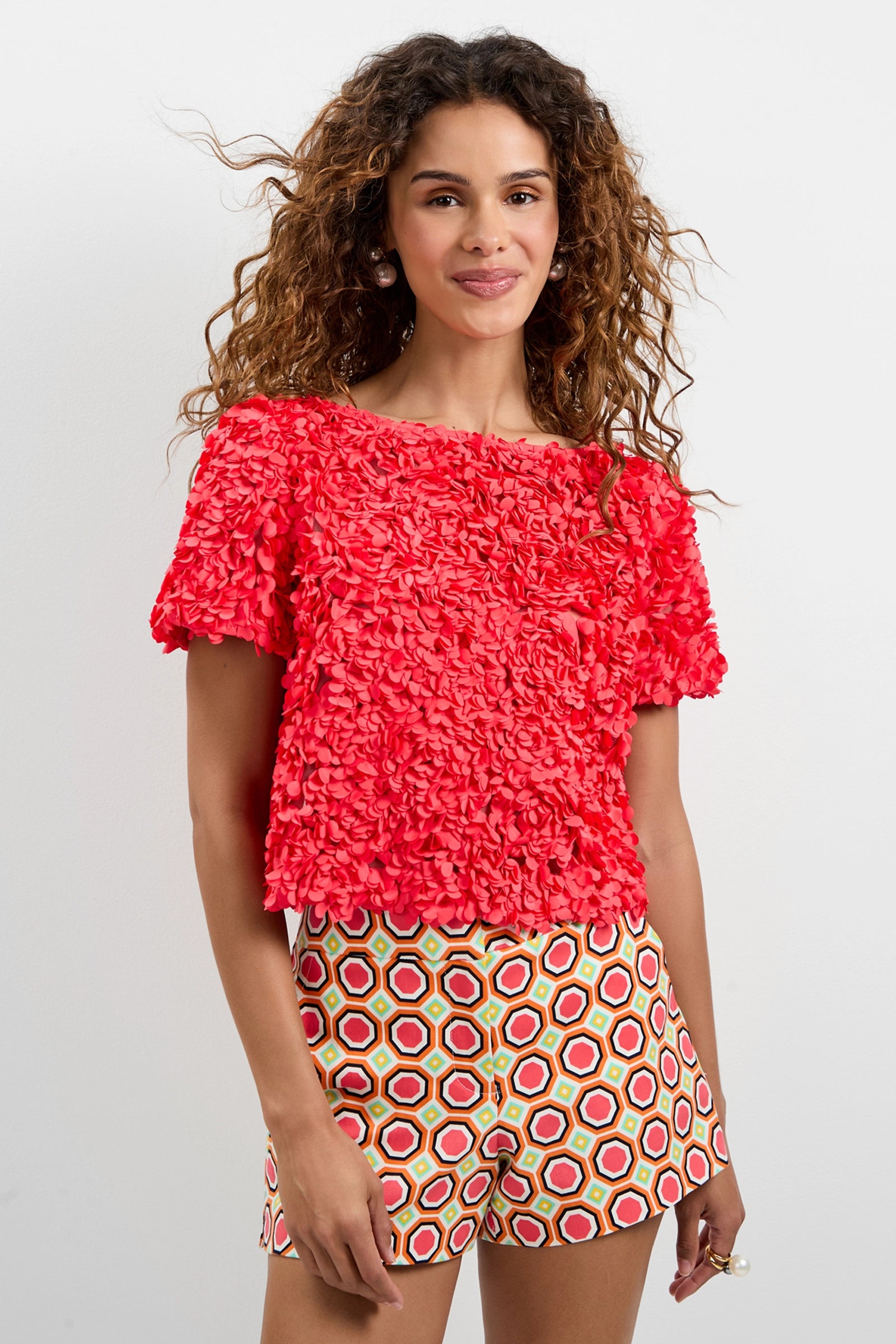 A woman with curly hair wears the Verdi 3D Floral Top, a bright red textured short-sleeve shirt, and patterned hexagonal shorts in red, yellow, and white, standing against a plain white background.