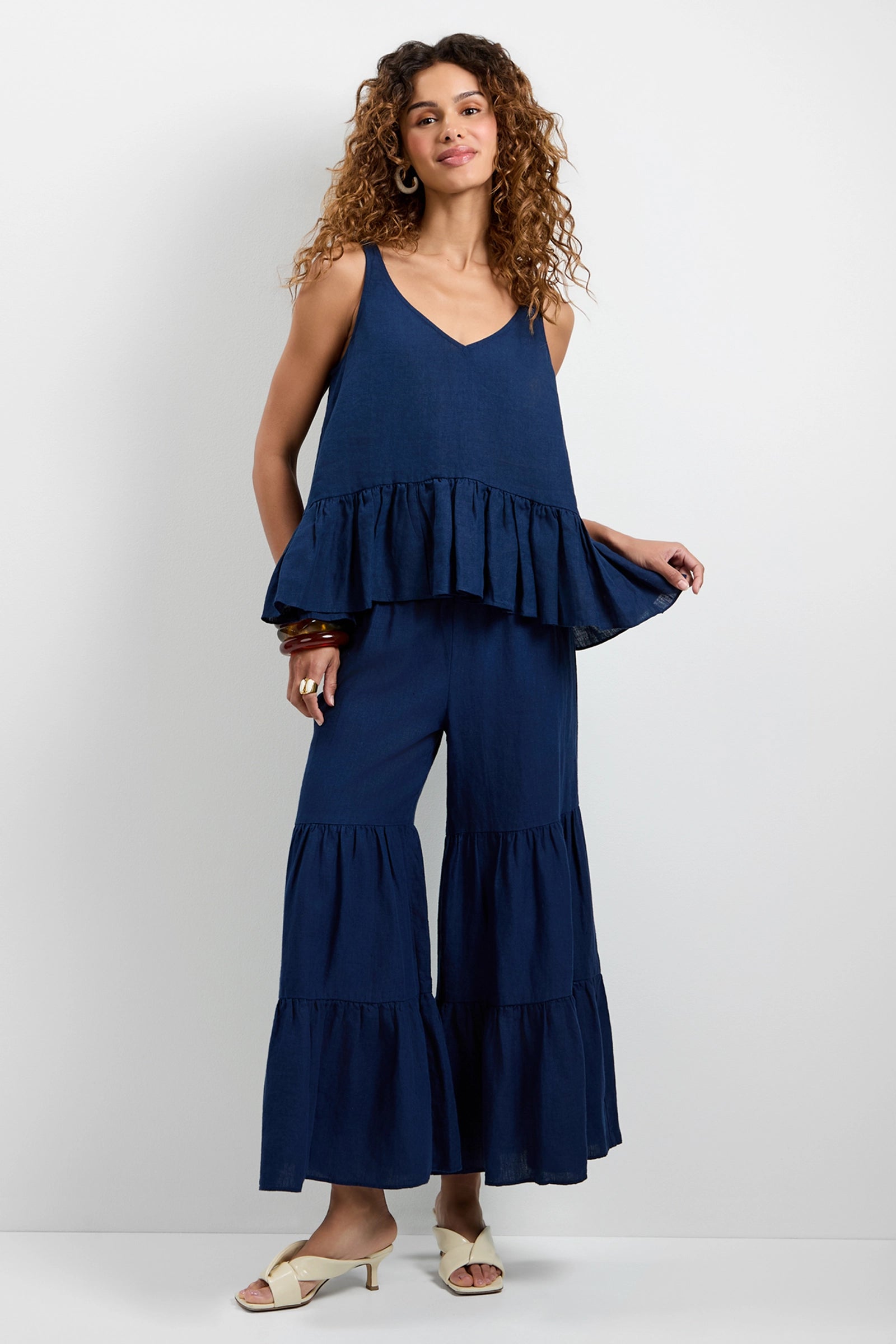 A woman with curly hair smiles against a white background, wearing the Bosco Tiered Ruffle Top— a loose-fitting, sleeveless navy top with a ruffled hem—paired with matching navy wide-leg pants and white heeled sandals.