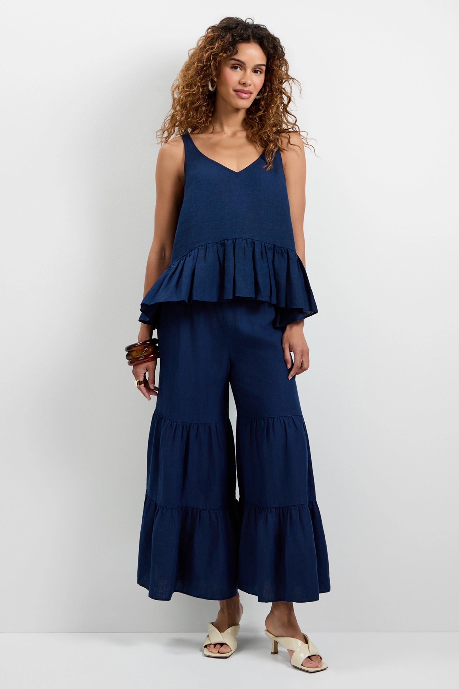 A woman with curly hair models the Chill Out Tiered Wide-Leg Pants in navy blue, paired with a matching sleeveless ruffled hem top. She accessorizes with bangles and cream open-toe heels against a plain background.