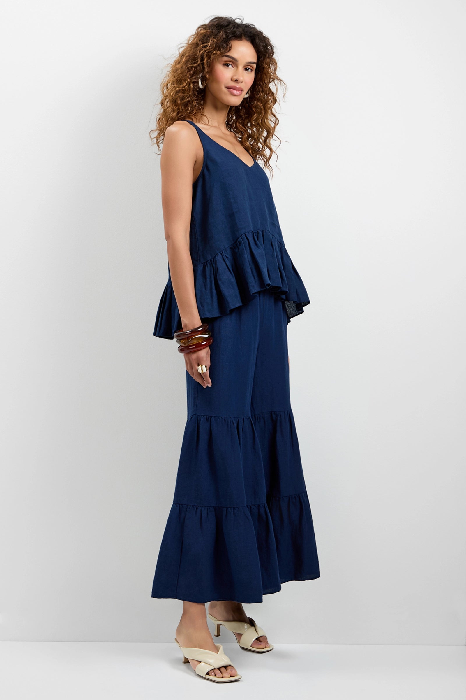 A woman with curly hair models the Bosco Tiered Ruffle Top in navy, paired with a matching tiered maxi skirt. She stands sideways, looking at the camera, accessorized with bracelets and cream heeled sandals against a plain white background.