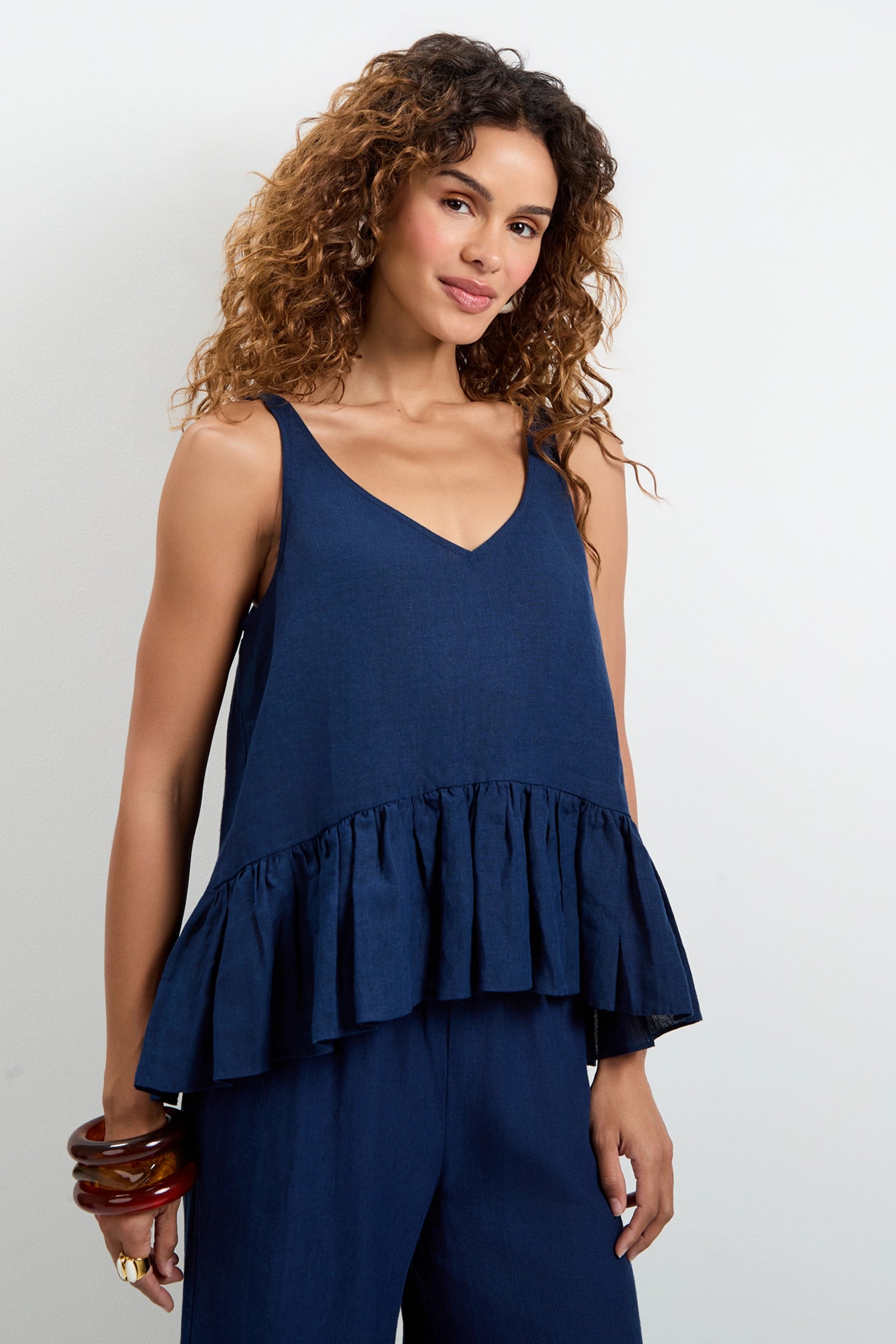 A woman with curly hair wears the Bosco Tiered Ruffle Top—sleeveless, navy blue, with a V-neck and ruffled hem—paired with matching wide-leg pants and chunky bracelets, standing against a plain light background.