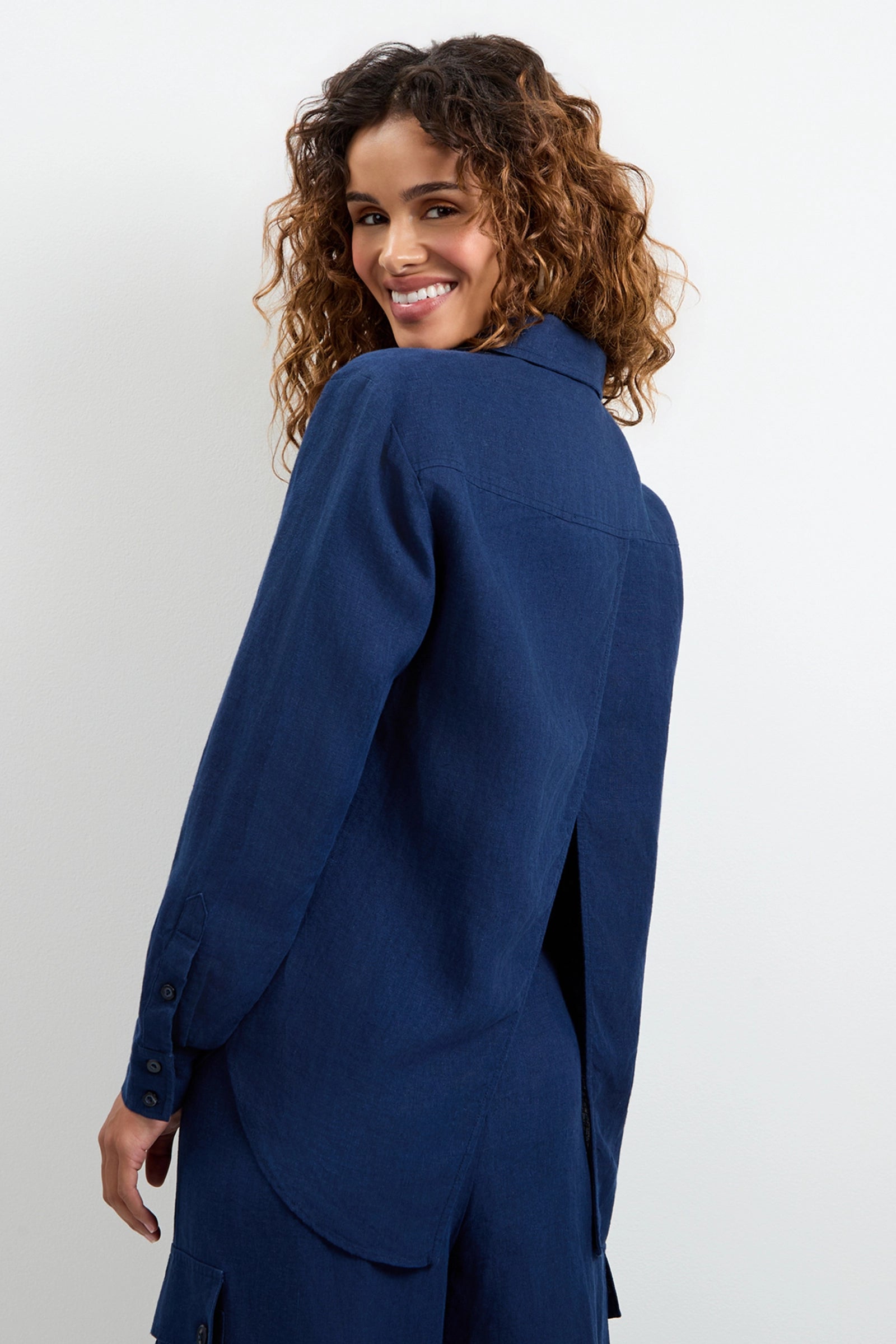 A woman with curly hair smiles over her shoulder, wearing the Barona Top—a navy blue long-sleeve shirt with a back slit—and matching pants, standing against a plain white background.