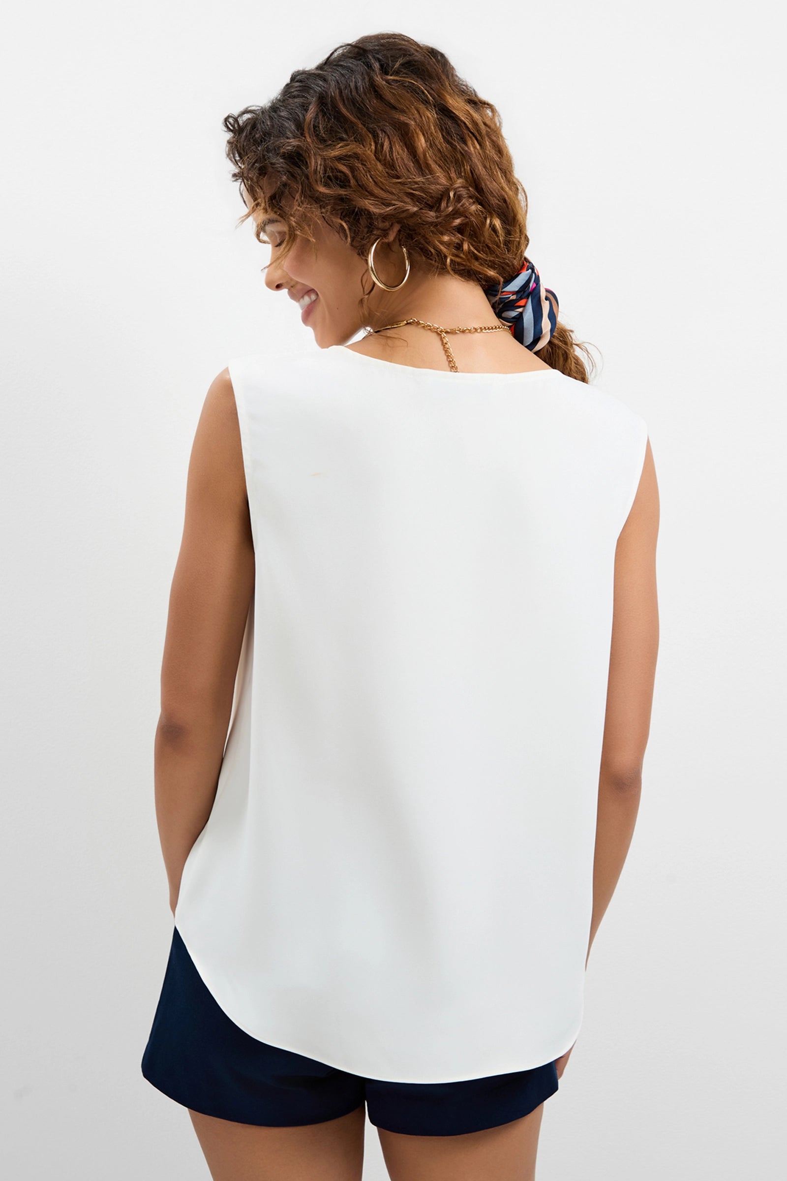 A woman with curly hair in a ponytail and hoop earrings stands with her back to the camera, wearing the sleeveless white Aprica Boatneck Top and dark shorts against a plain white background.