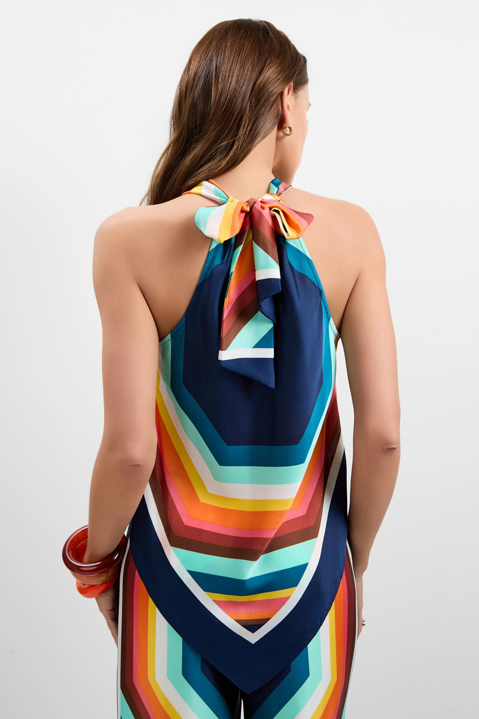 A woman with long brown hair, shown from the back, wears the Lavani Halter Neck Top and matching pants with bold geometric diagonal stripes in blue, orange, yellow, and pink. She accessorizes with large red bangles on her wrist.